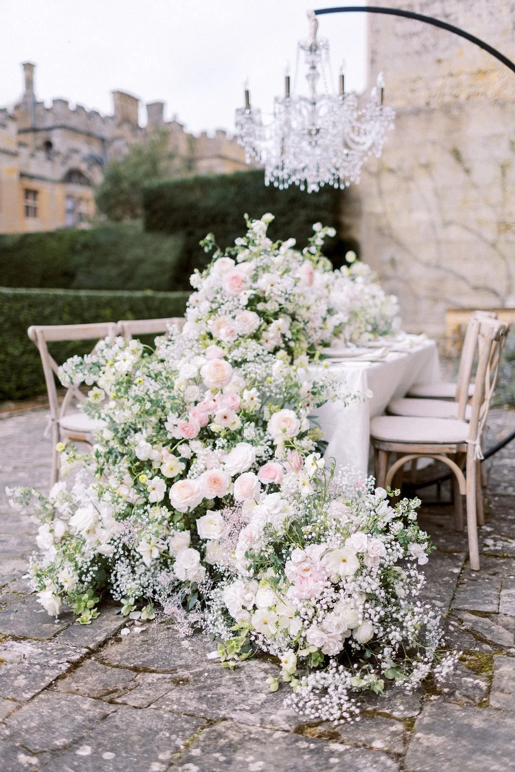 Elegant outdoor wedding reception table decorated with a lush arrangement of pink and white flowers, with a crystal chandelier hanging above, set on cobblestone ground with historic castle buildings in the background.
