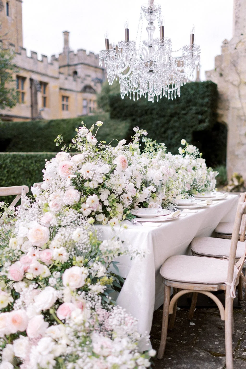 Elegant outdoor dining table decorated with large floral arrangements of white and blush pink flowers, set beneath a crystal chandelier with a castle-like stone building in the background.