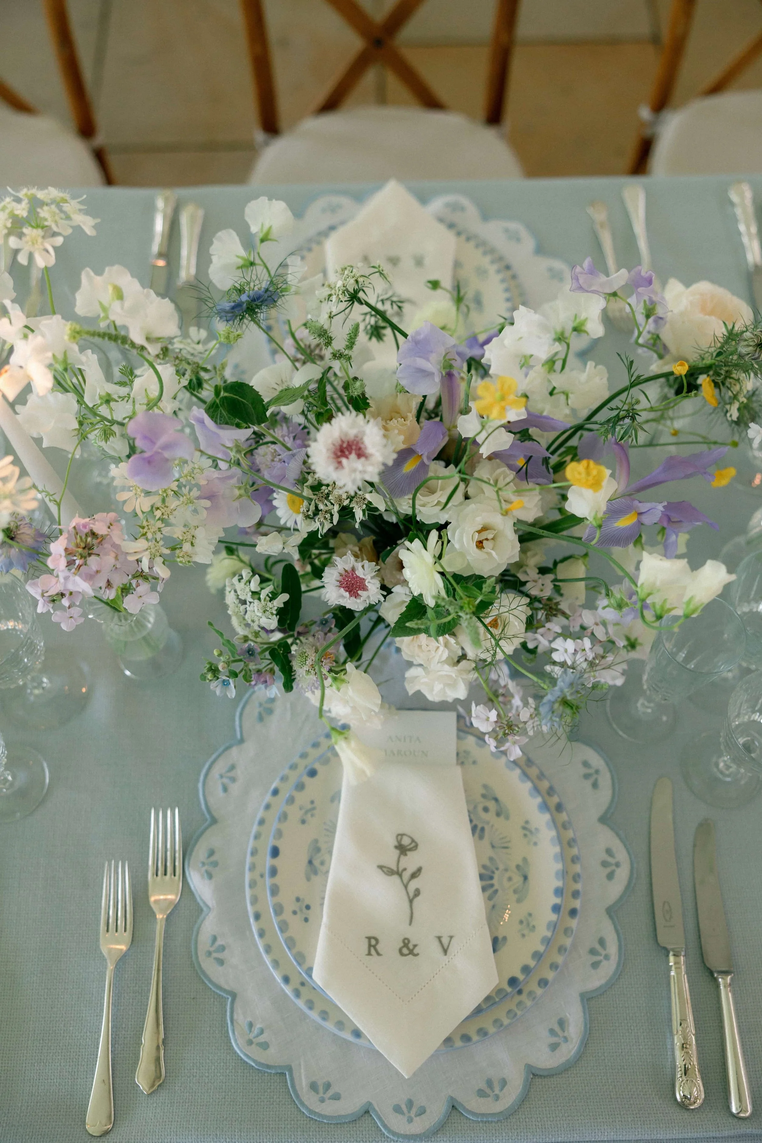 A table setting with a floral centerpiece, tableware, and a personalized napkin at a wedding or special event. Wilderness Reserve, Suffolk Florals By The Botany House