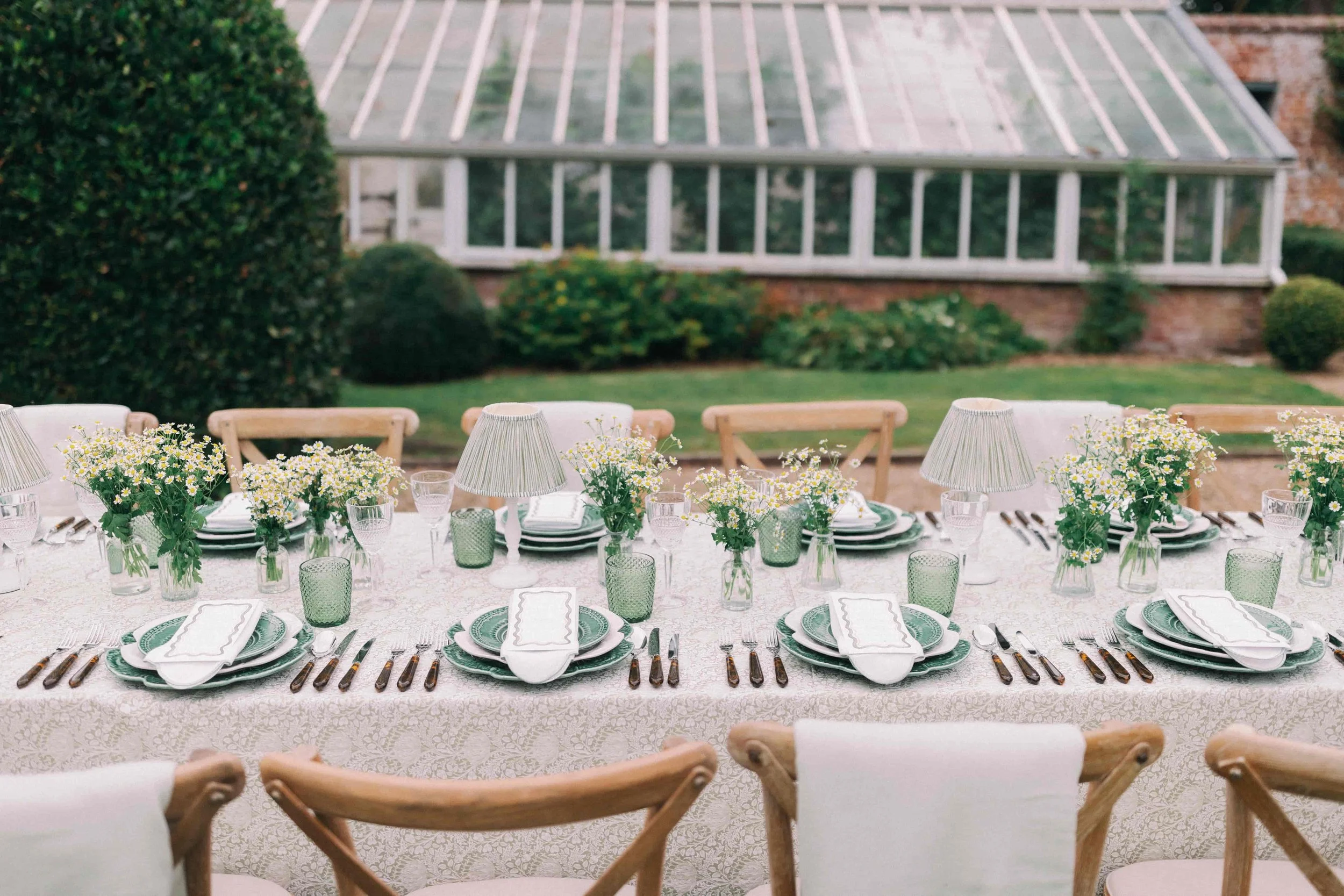 Outdoor dining table set with white tablecloth, green and white patterned plates, silver cutlery, white napkins, glassware, small lamps, and floral centerpieces of daisies in glass vases, with a garden and greenhouse in the background.