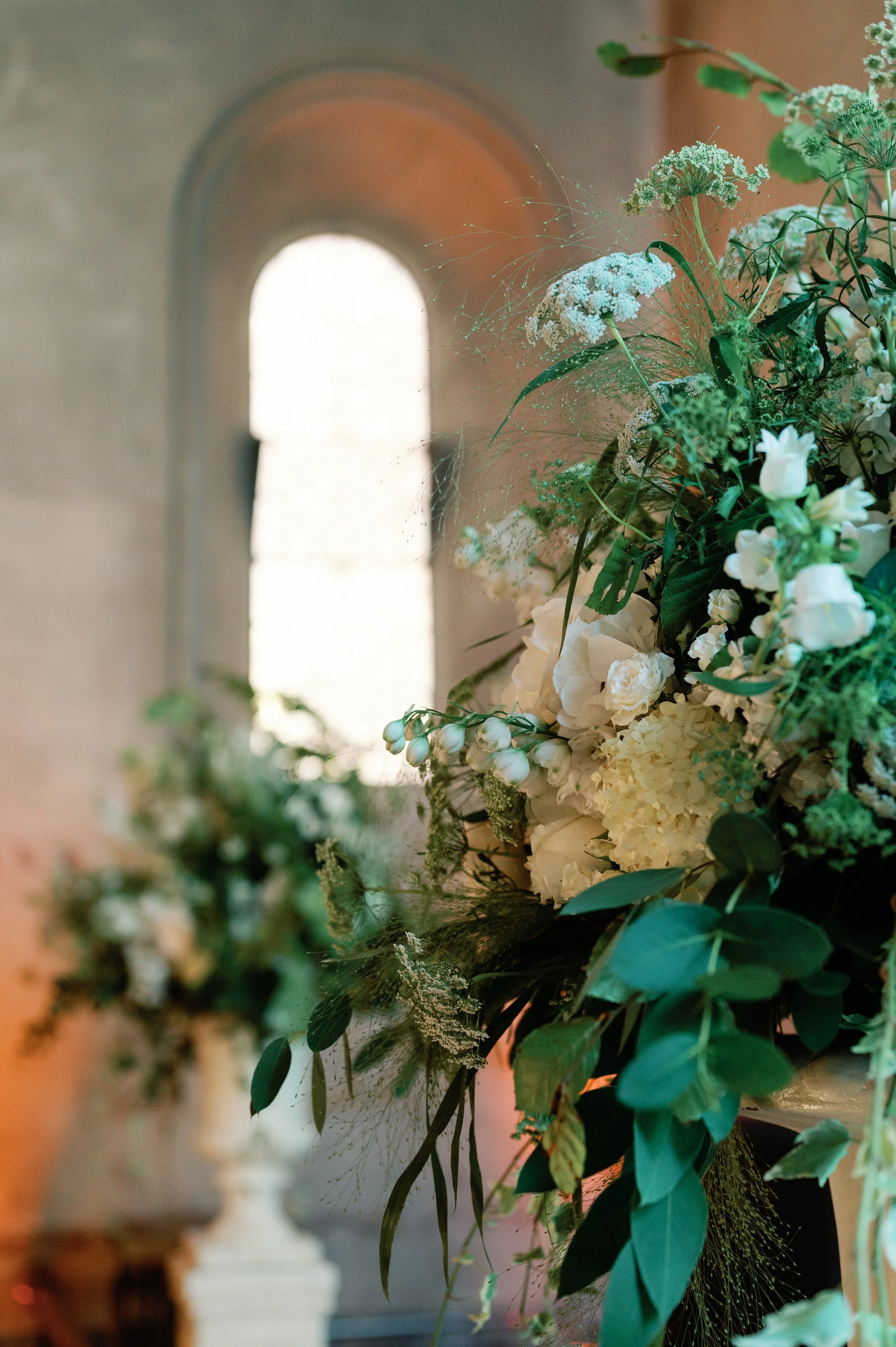 White and green floral arrangement with roses, hydrangeas, and greenery inside a building with a window in the background.