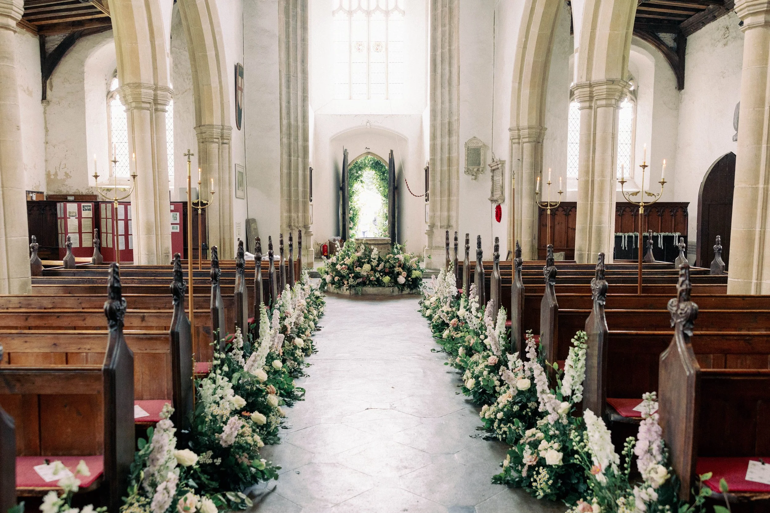 Interior of a church decorated with white and pink floral arrangements along the aisle, with wooden pews on either side and a large floral display at the altar, lit by chandeliers and natural light. A countryside wedding in Norfolk