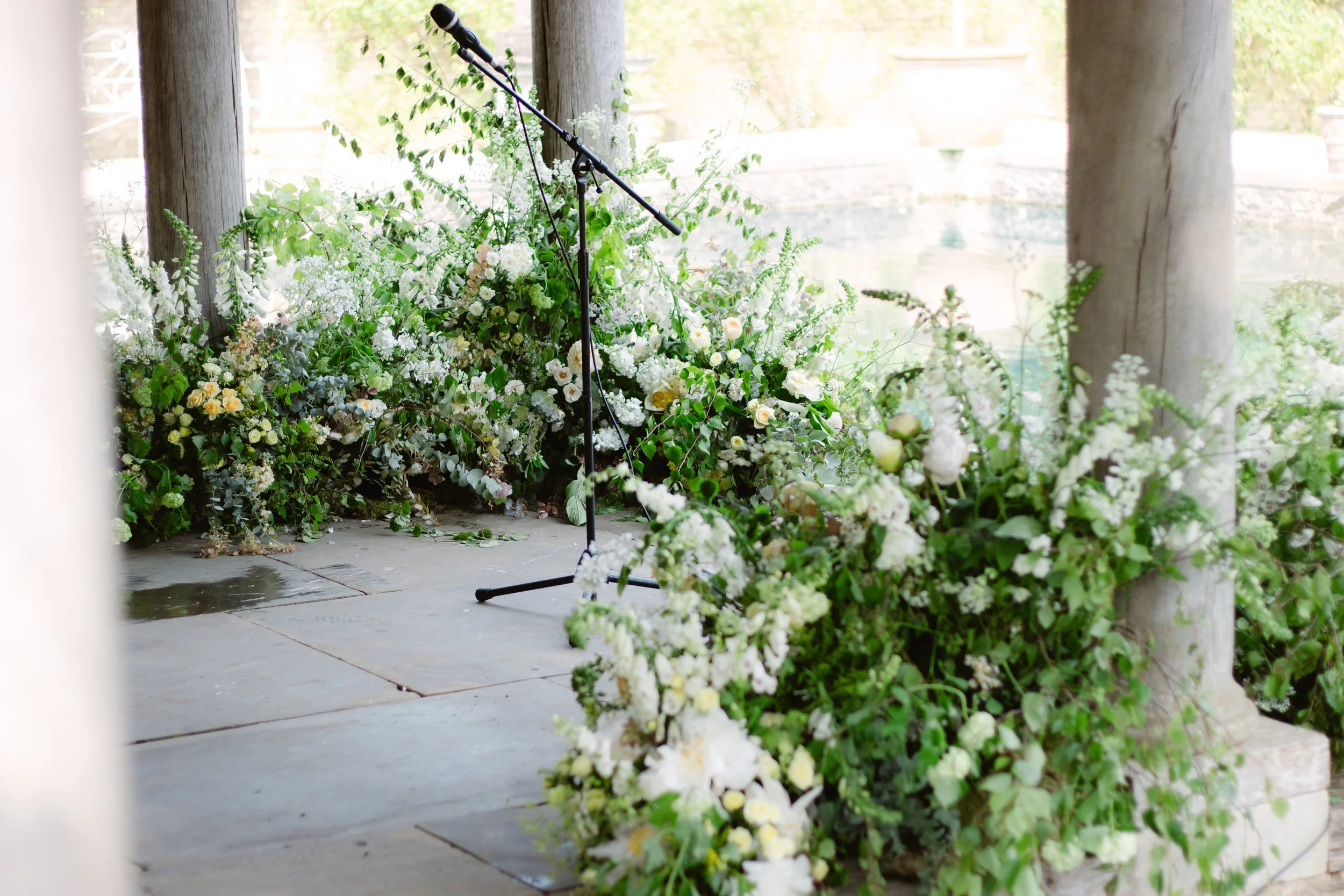 A microphone on a stand surrounded by white and light-colored flowers on a stone surface, with a background of wooden columns and a pond.
