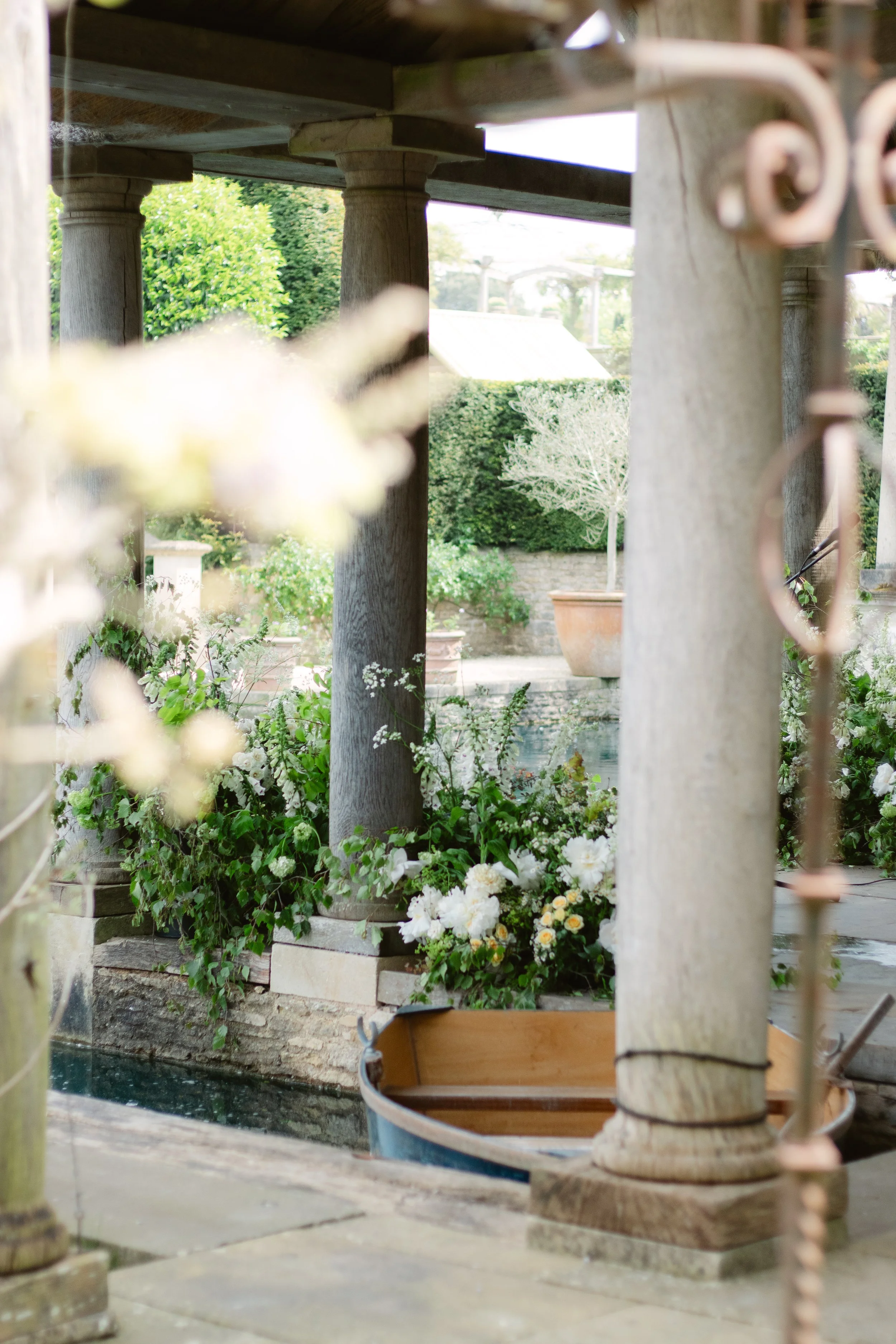A garden scene featuring stone columns, lush green plants, and a small boat docked at a stone edge, with flowers and hedges in the background.