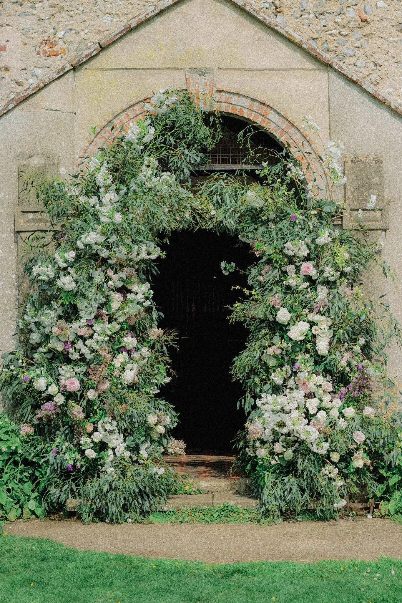 Flower archway with greenery and white and pink flowers outside a stone building entrance.