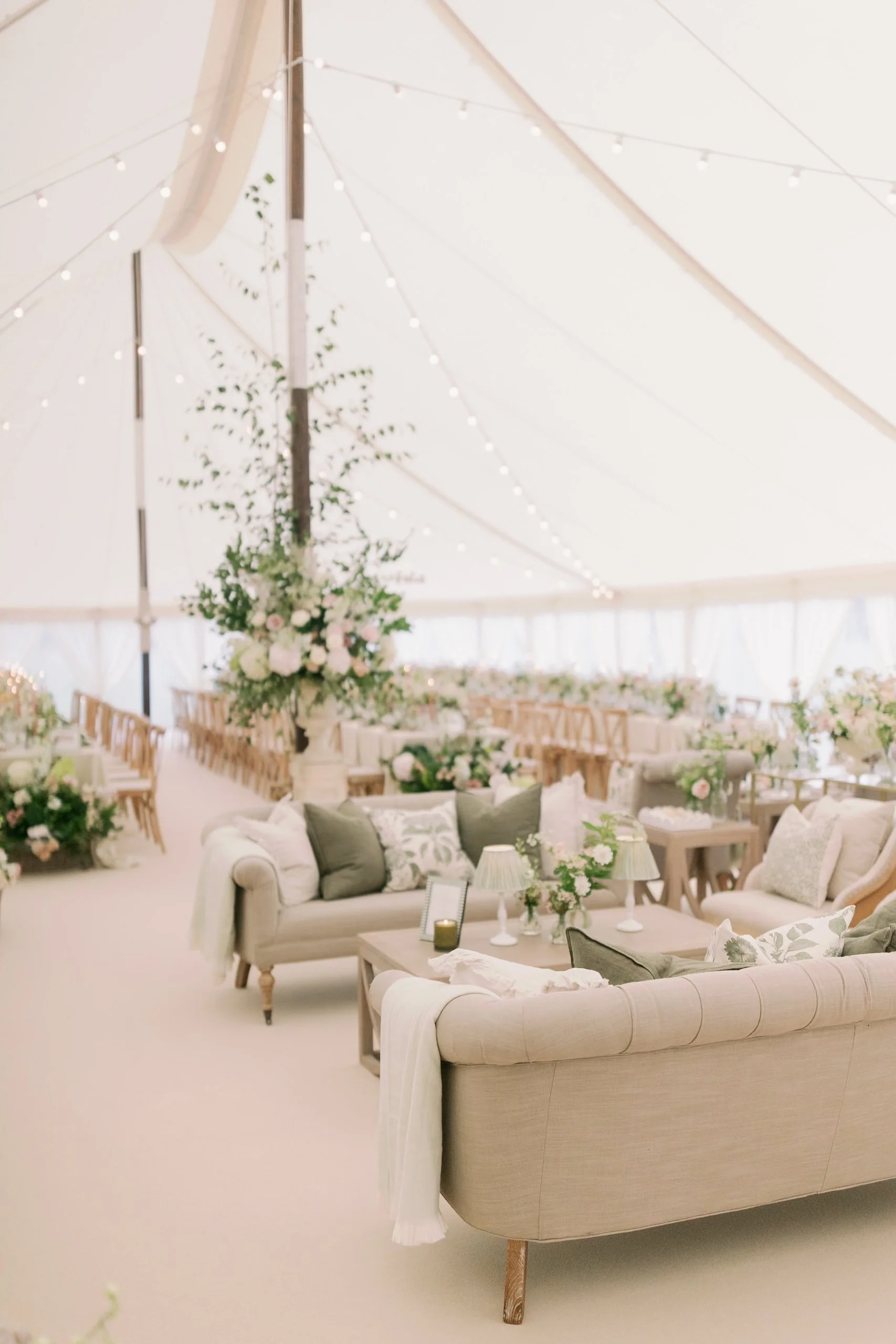 Elegant wedding reception inside a large, white sailcloth marquee decorated with string lights and floral arrangements. There are multiple tables with floral centerpieces, surrounded by wooden chairs, and cozy sofas with decorative pillows in neutral