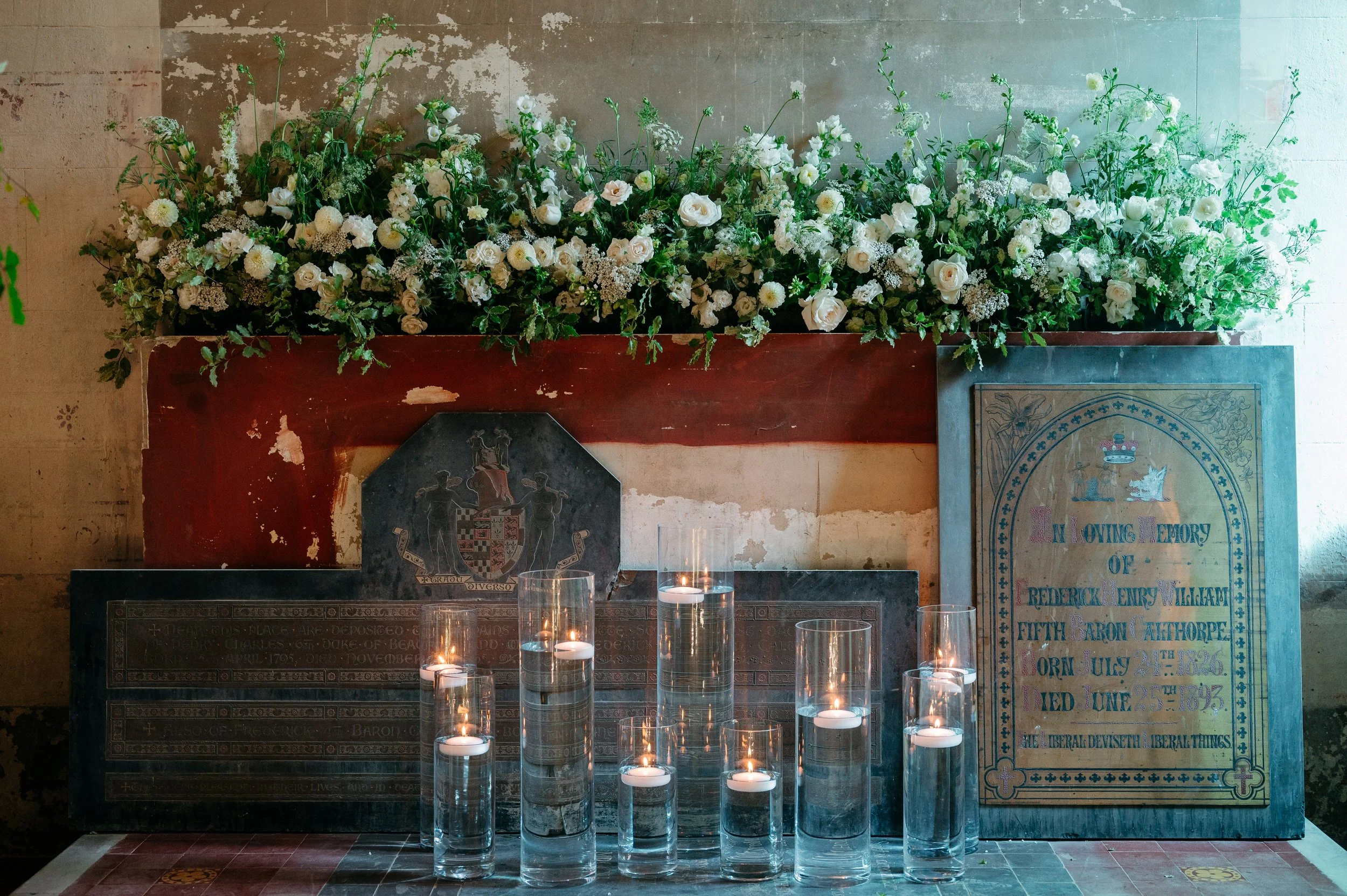 A memorial altar with a large arrangement of white flowers, several lit candles in glass holders, and memorial plaques, including one honoring Frederick Henry William, made of stone and metal, with a coat of arms and an inscription.