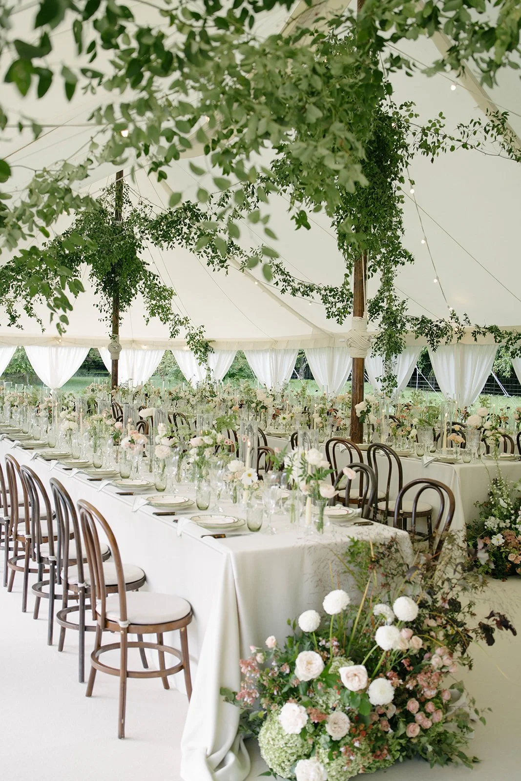A beautifully decorated outdoor wedding reception area under a large white marquee tent, adorned with hanging greenery and string lights. Long tables with white tablecloths are set with plates, glasses, and floral centerpieces, and there are wooden c