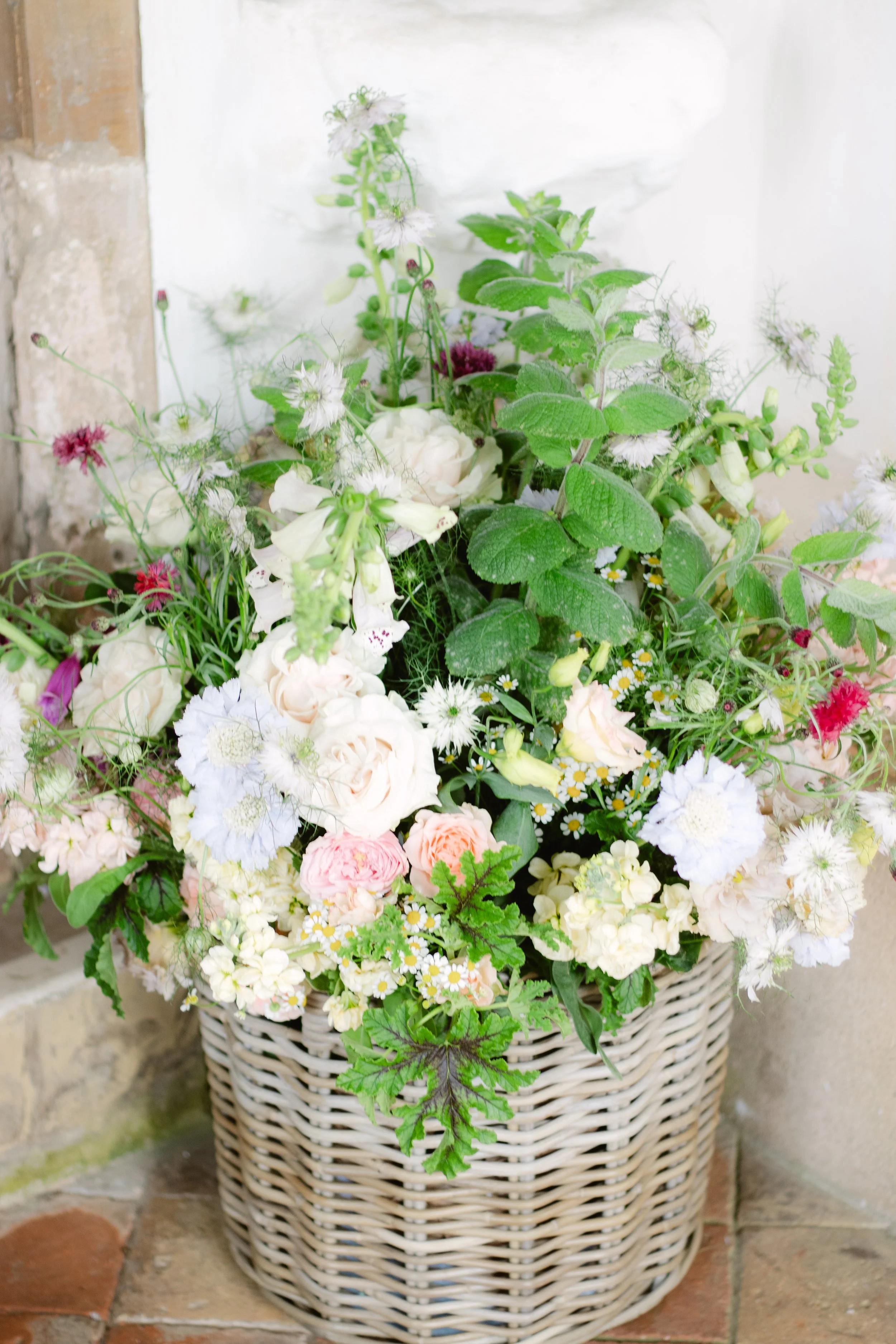 A basket filled with various light-colored and pastel flowers, including roses, daisies, and other blossoms, rests on a brick surface against a wall.