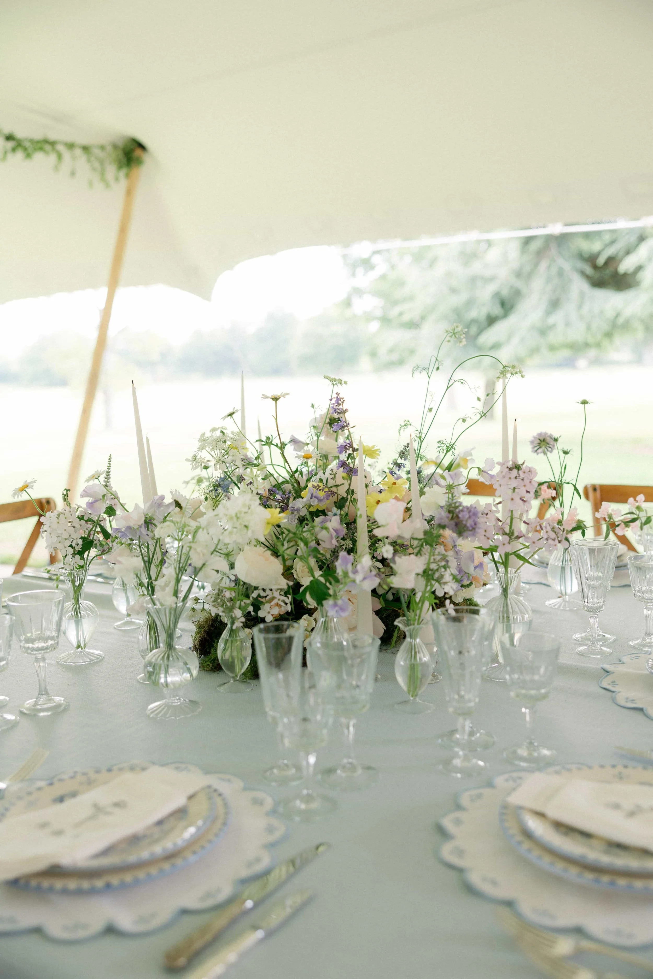 A round table set for a formal event with a centerpiece of light-colored wildflowers in glass vases, surrounded by glassware and place settings with scalloped-edged plates and cloth napkins, in a white marquee. The Botany House Florist at Wilderness
