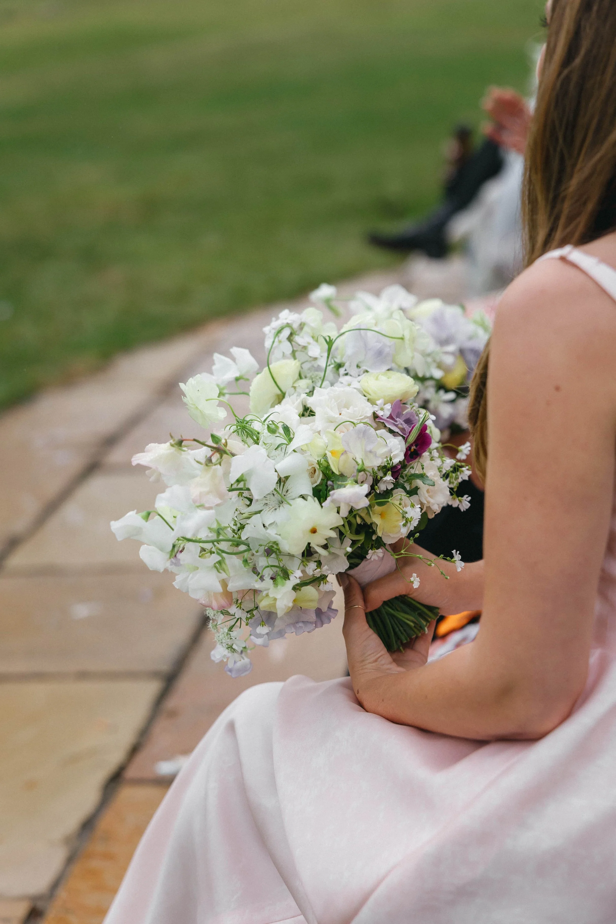 A bridesmaid wearing a pink dress holding a bouquet of white and light purple flowers at a wedding, with other people seated in the background. Wilderness Reserve, Suffolk Florals By The Botany House