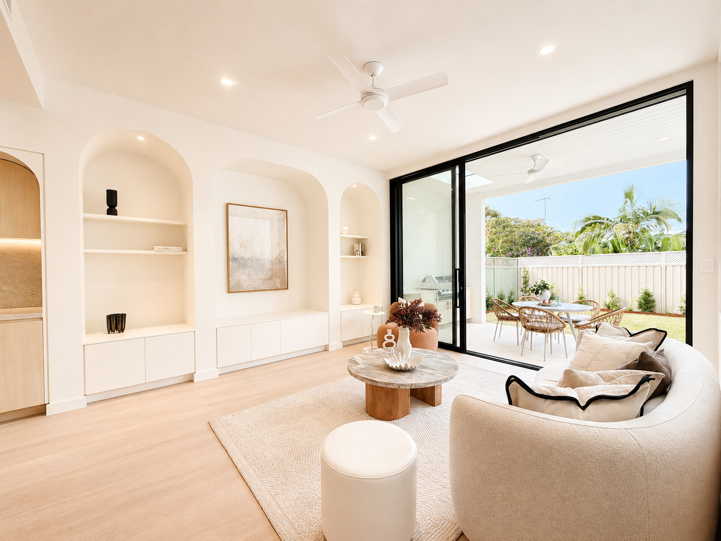Bright living room with beige sofa, round wooden coffee table, and sliding glass door opening to a backyard patio with outdoor dining furniture and lush greenery.