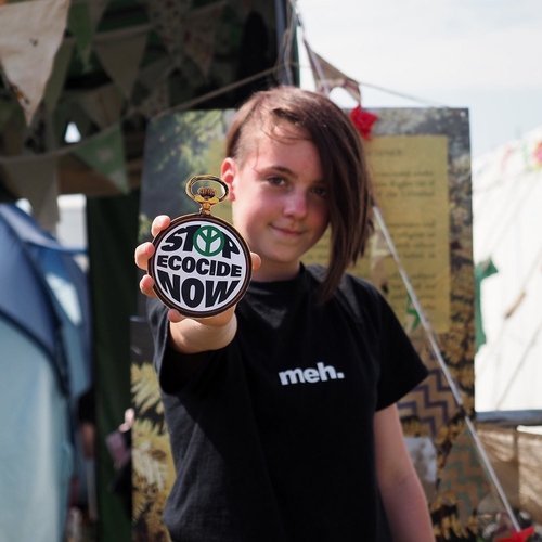 niña sujetando reloj que dice 'Stop Ecocide now' delante de un puesto de la campaña en un festival de verano