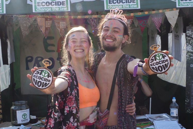 pareja sujetando un reloj que dice 'Stop Ecocide now' delante de un puesto de la campaña en un festival de verano