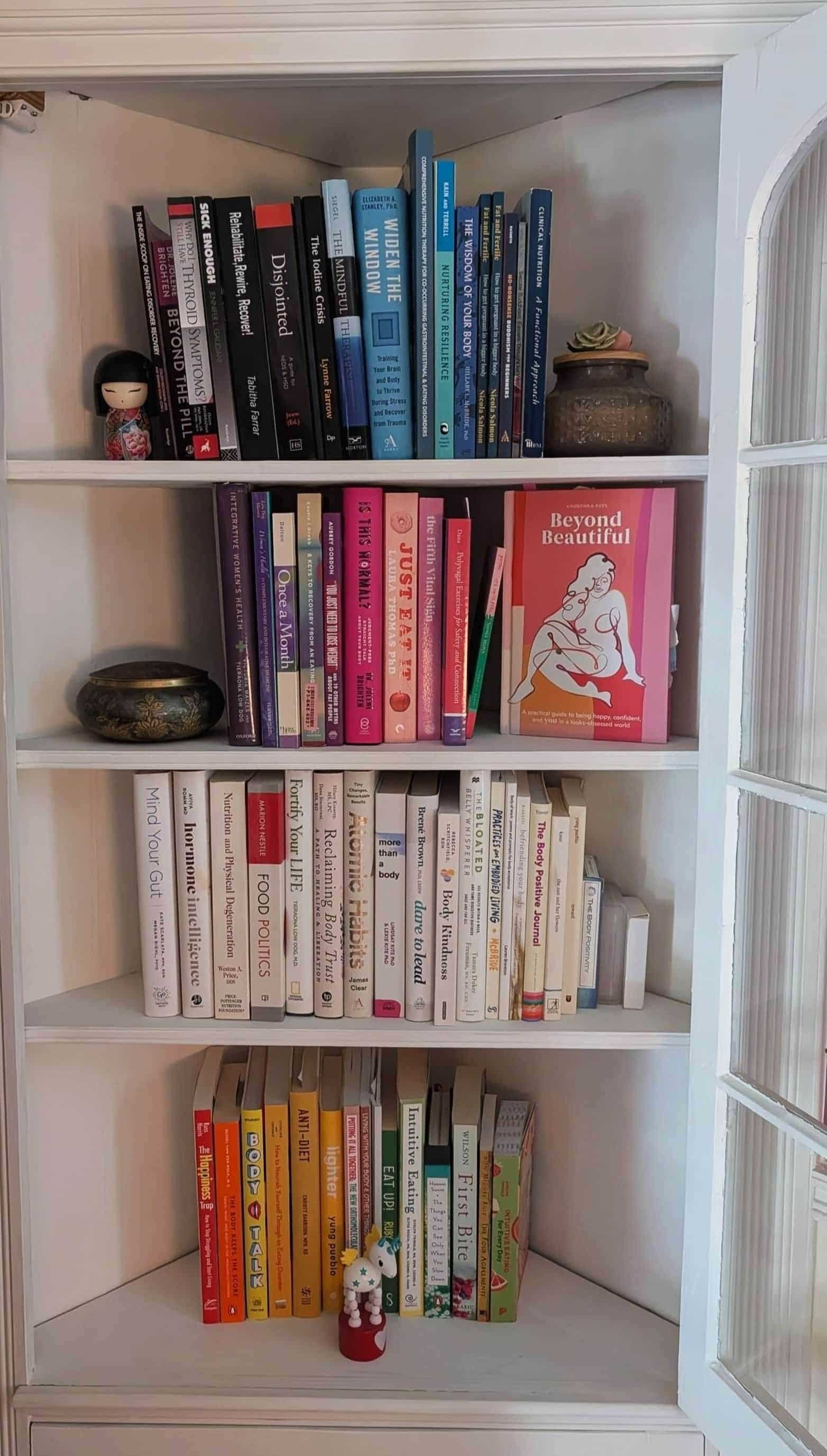 A neatly organized bookshelf with colorful books, figurines, and decor—plus a touch of weight-inclusive nutrition inspiration—by a glass-paned door.
