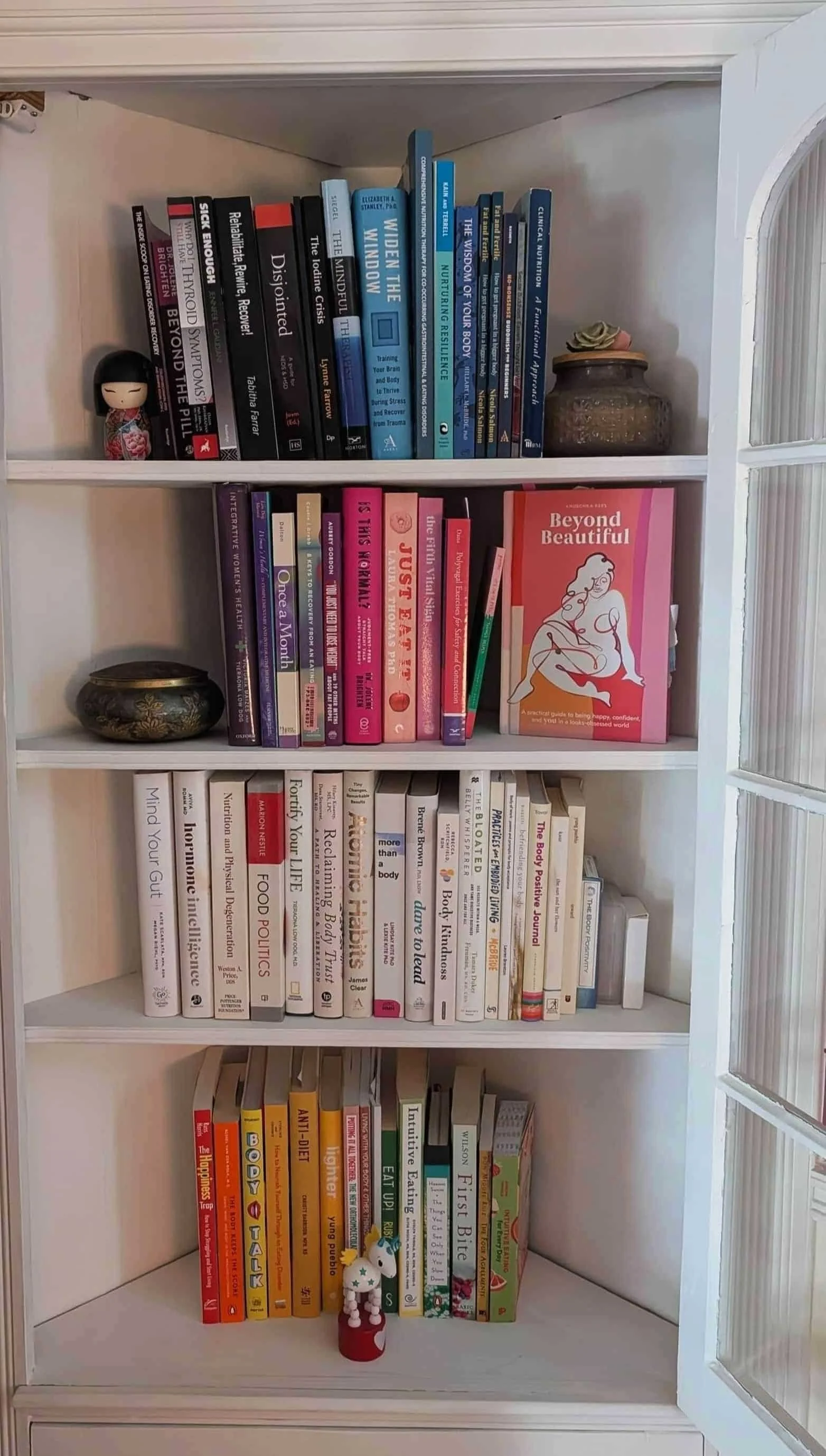 A neatly organized bookshelf with colorful books, figurines, and decor—plus a touch of weight-inclusive nutrition inspiration—by a glass-paned door.