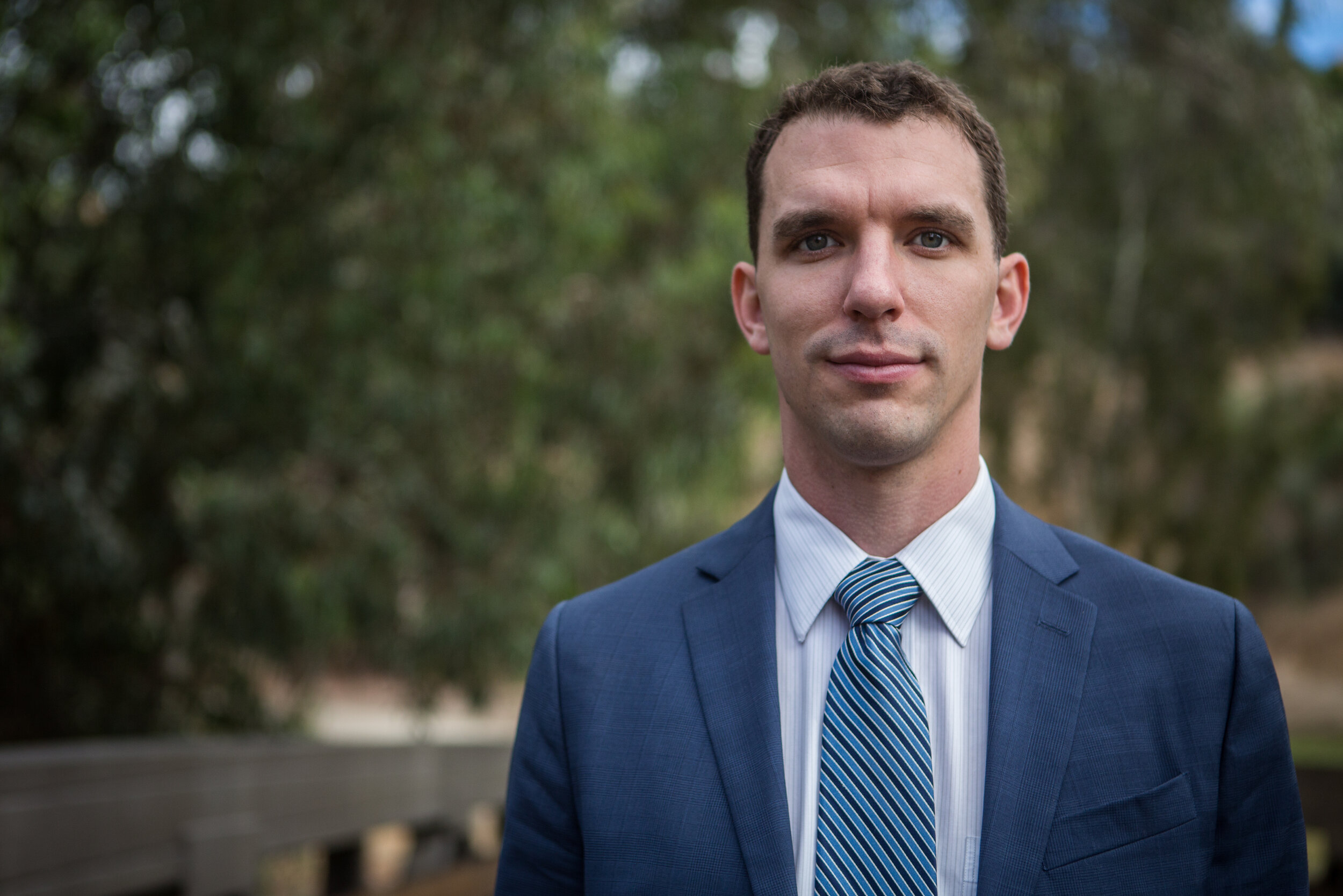 A man in a dark blue suit and light blue striped tie standing outdoors with trees in the background.
