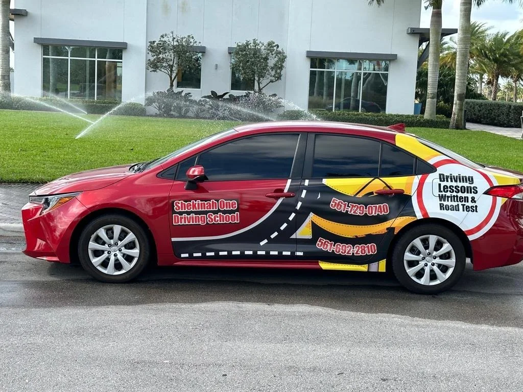 Red car with advertising for Shekinah One Driving School, featuring graphics of a road, route, and phrases like 'Driving Lessons Written & Road Test' along with contact numbers 561-729-0740 and 561-692-2810.