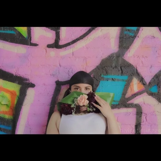 Person holding flowers in front of their face, standing against a colorful graffiti wall.