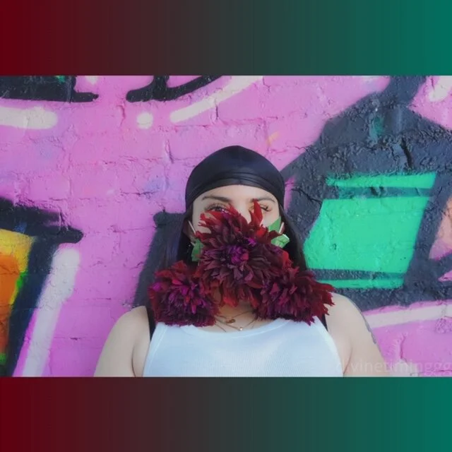 Person with flowers covering face, standing against colorful graffiti wall