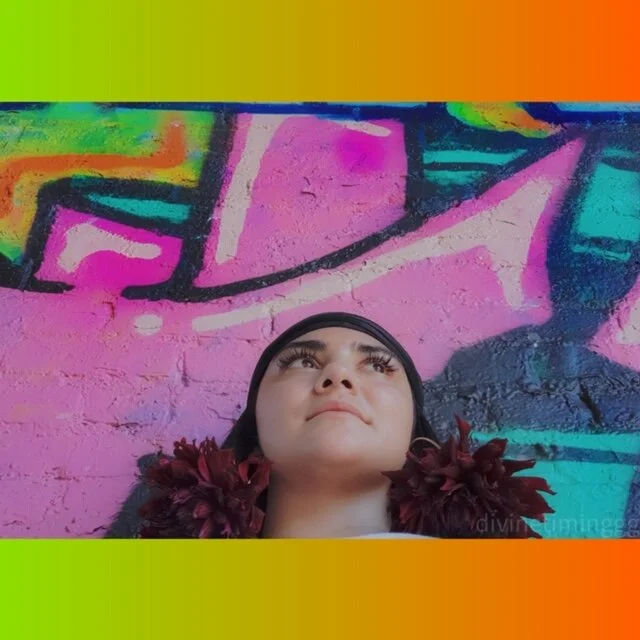 Person looking up in front of colorful graffiti wall, wearing a headscarf and a floral accessory.