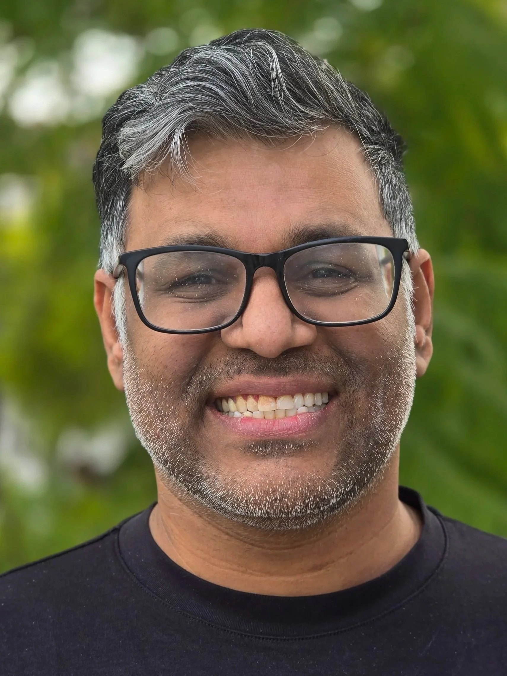 A smiling man wearing black glasses and a black T-shirt posing for a photo in front of a green tree