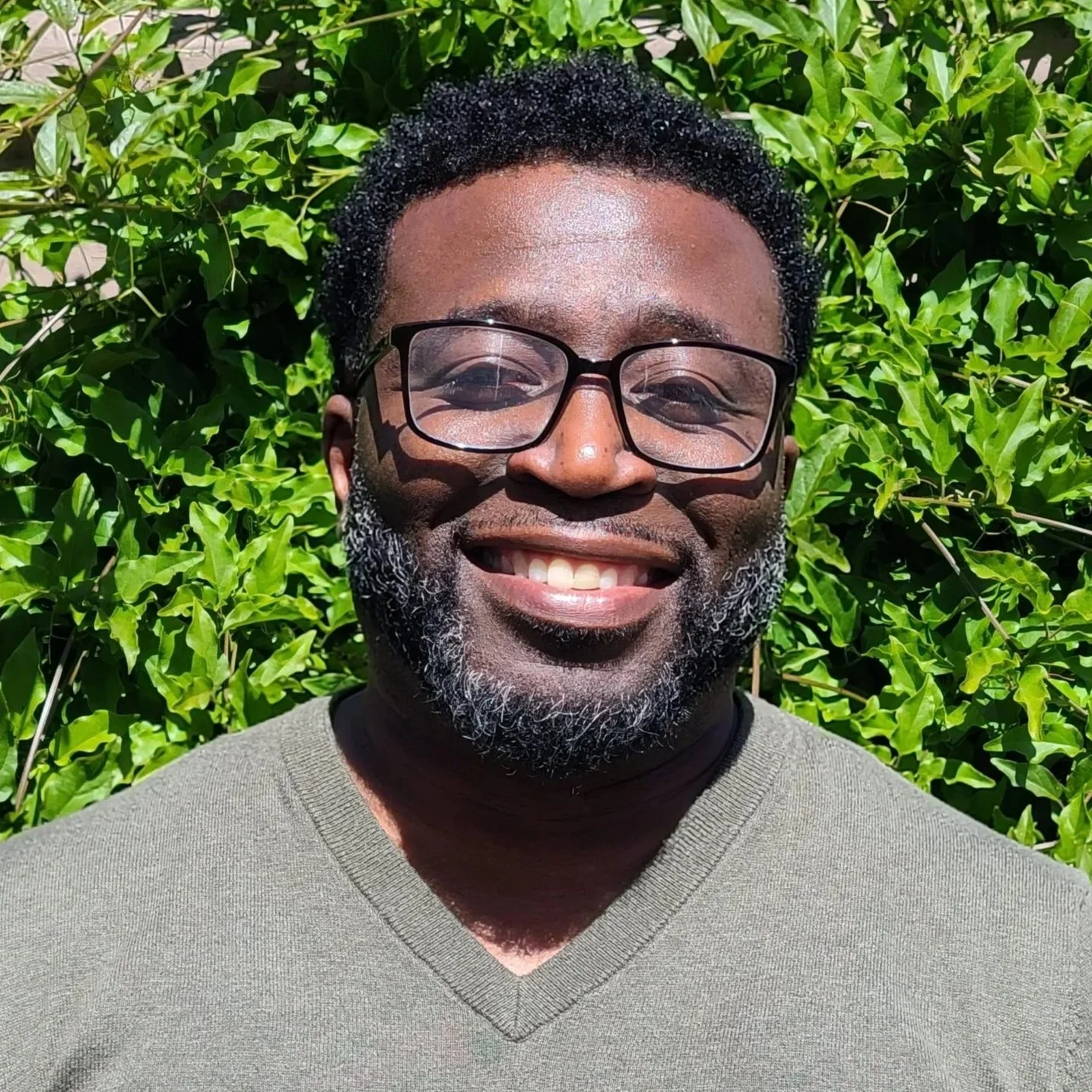 A smilng man wearing black-rim eyeglasses standing in front of green shrubbery on a sunny day
