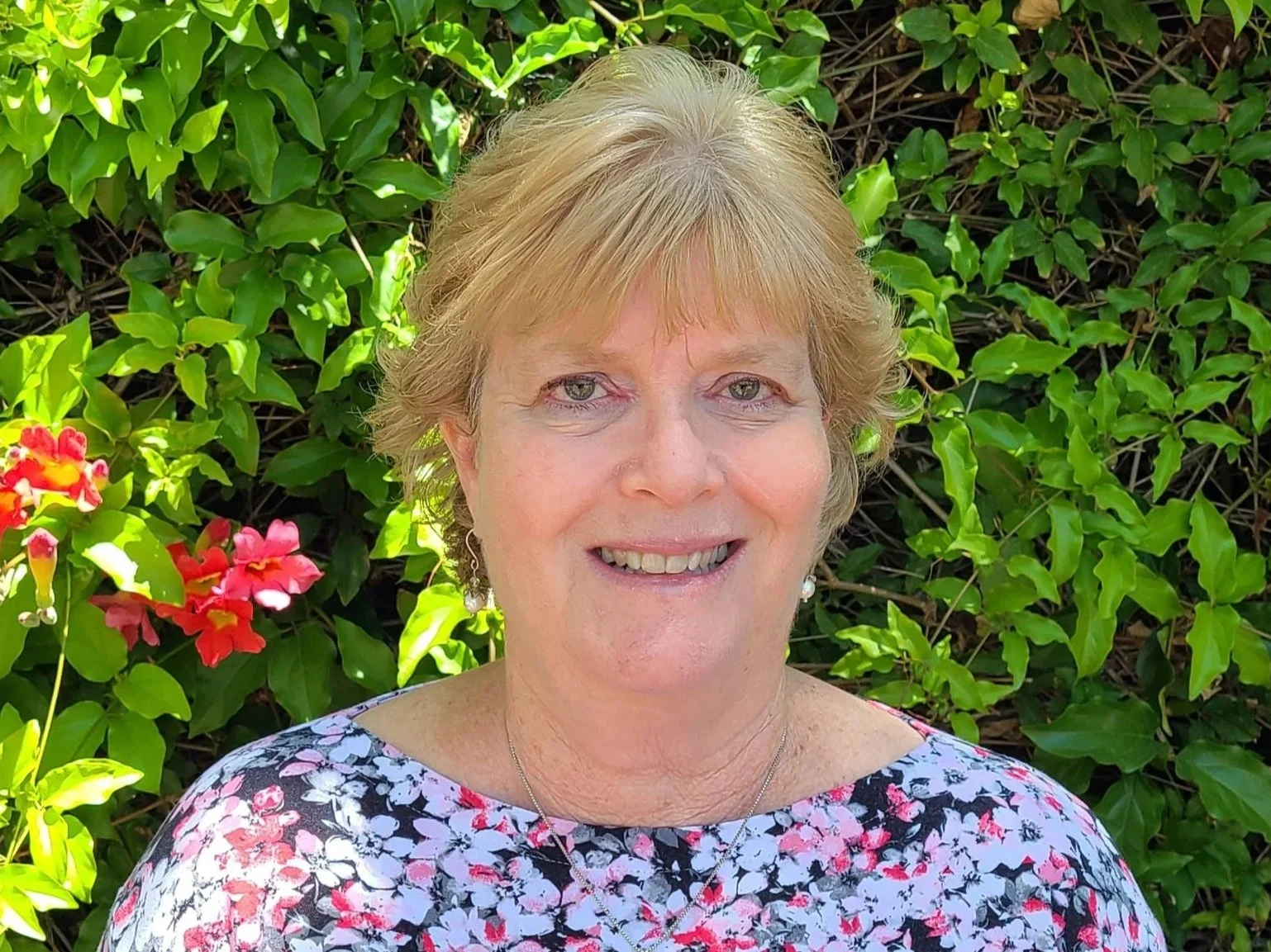 A woman in a floral blouse standing in front of shrubbery on a sunny day