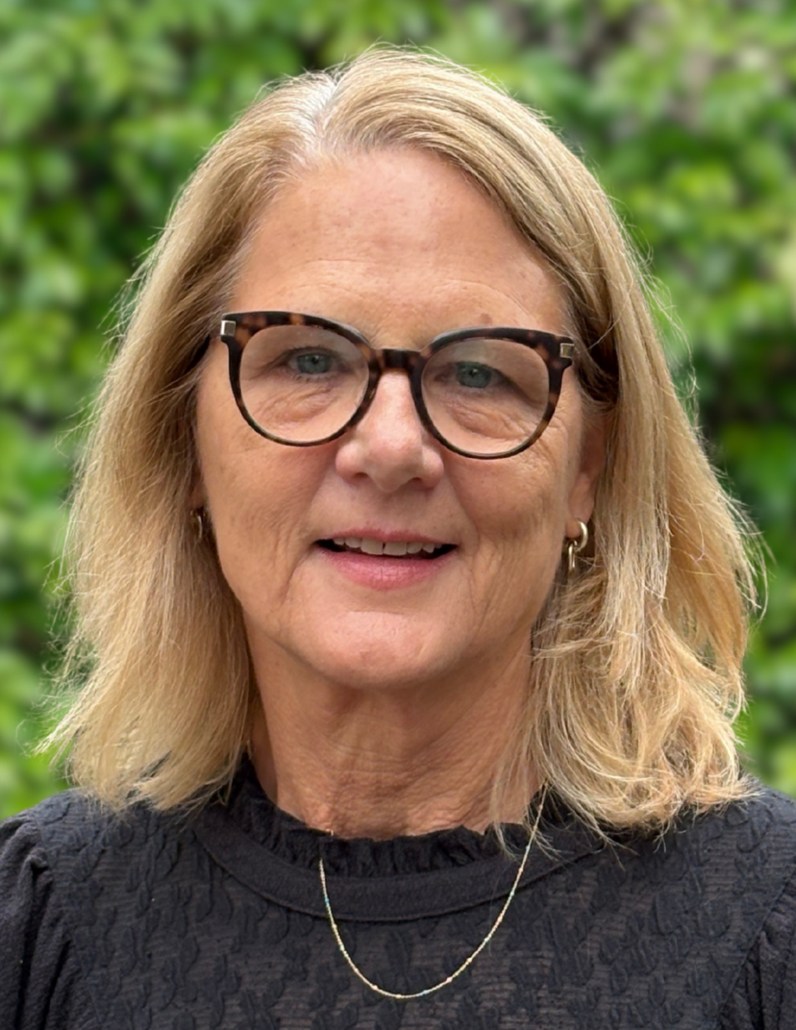 A woman wearing eyeglasses, a necklace, and a dark blouse posing for a photo outside in front of a green tree
