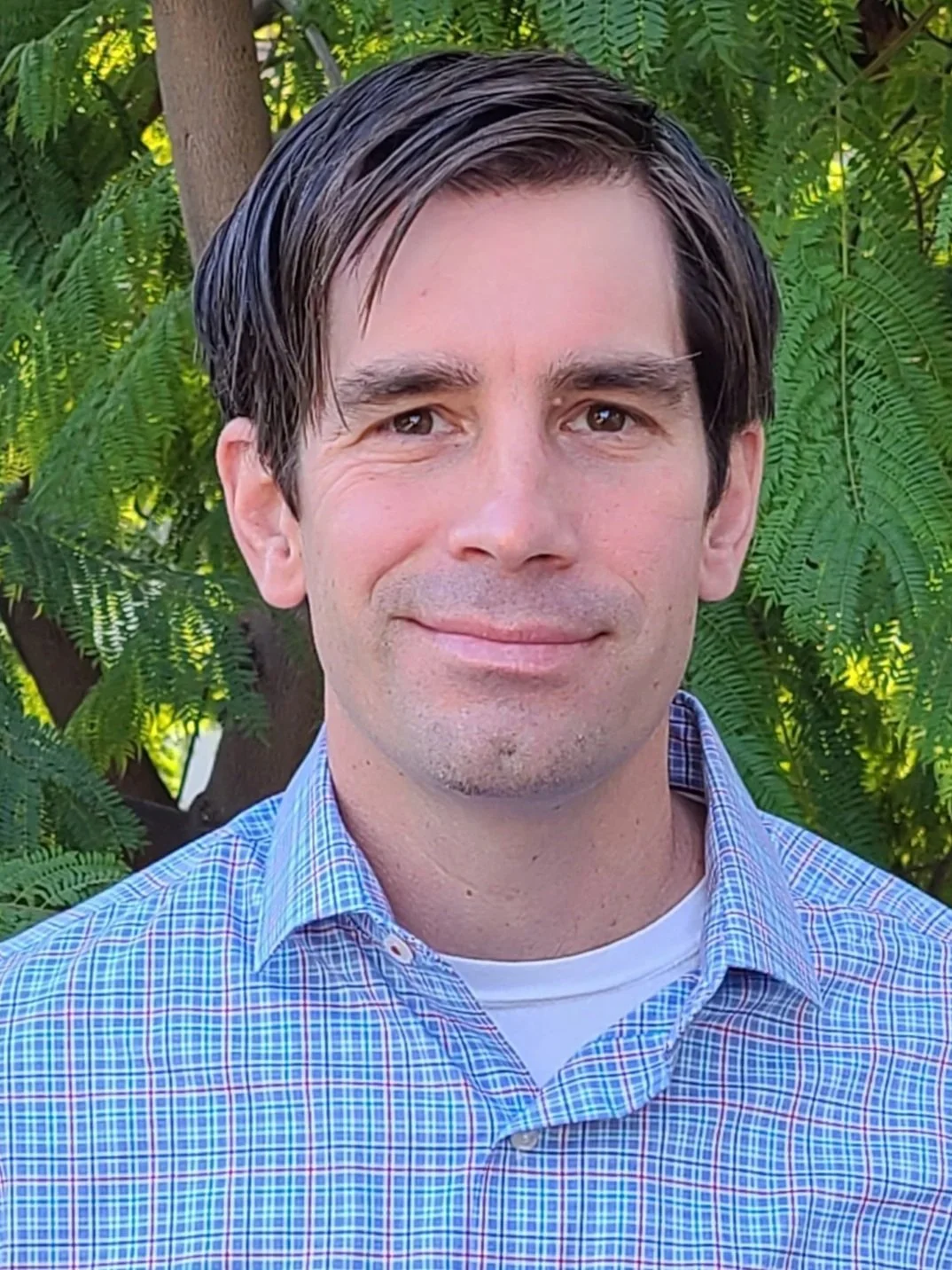 A man wearing a blue dress shirt standing in front of green shrubbery on a sunny day