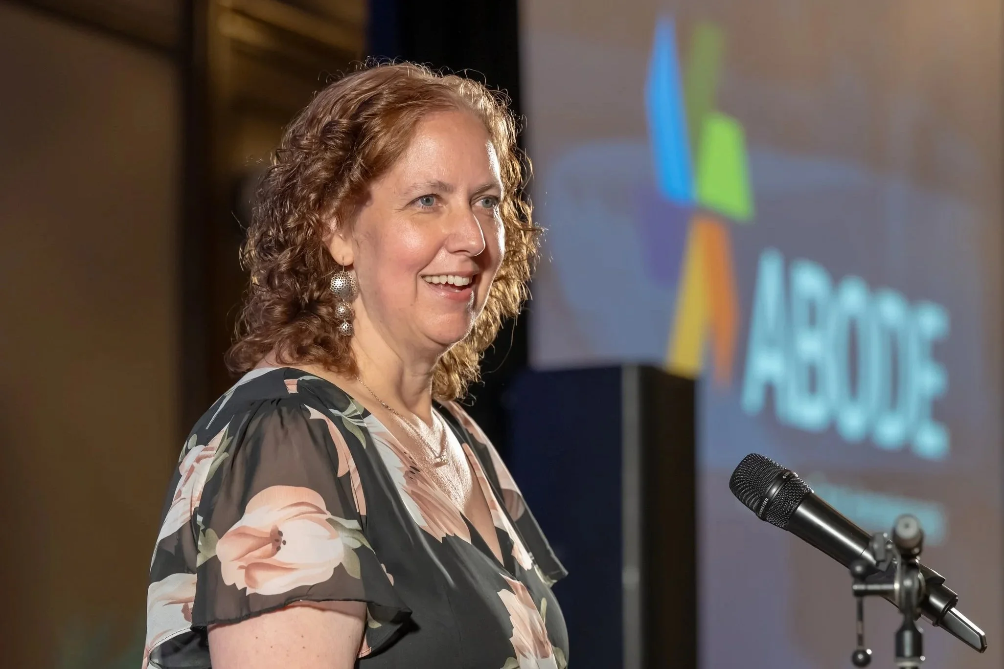 A smiling woman in a floral dress delivers a speech at a fundraiser in a hotel ballroom