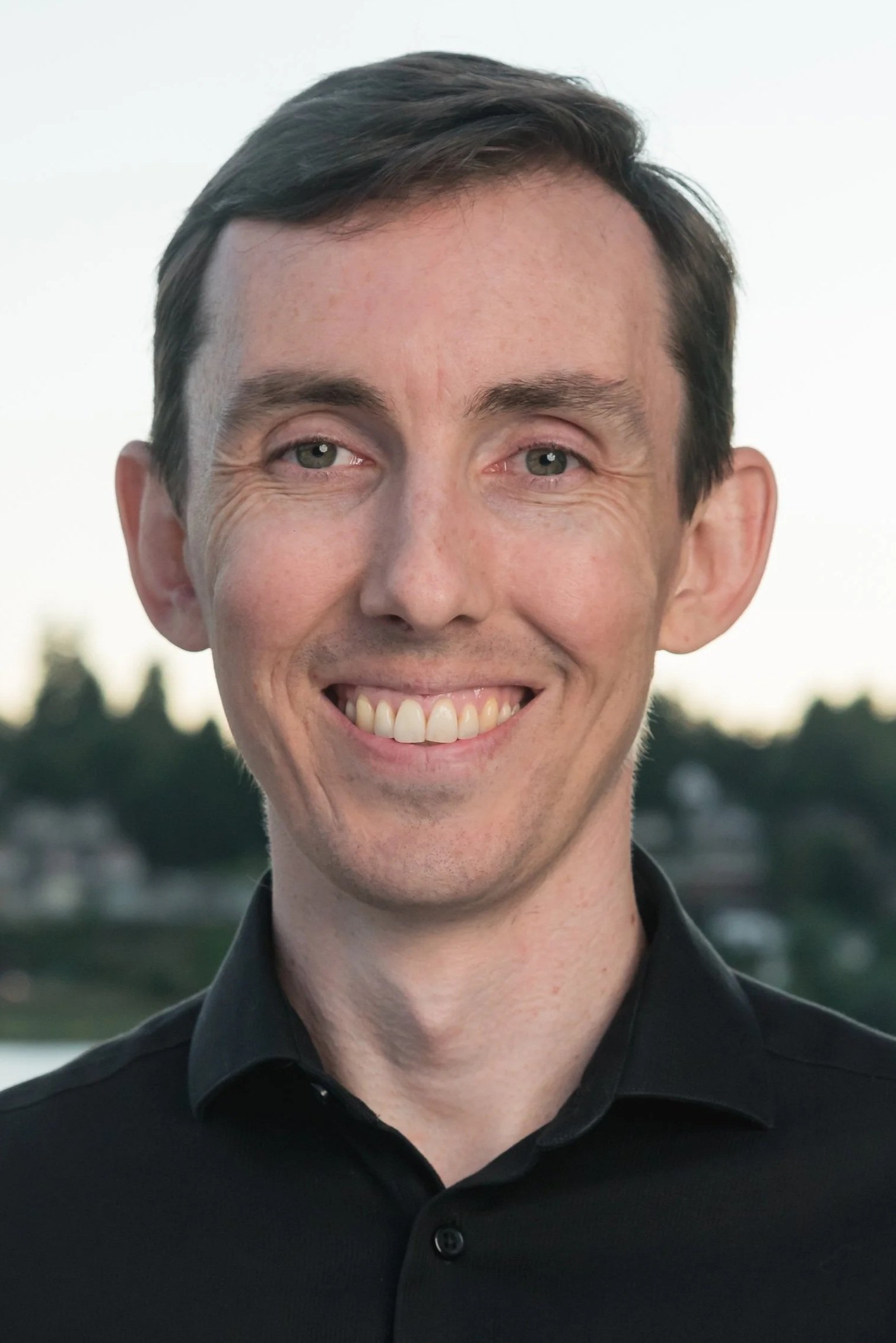 A smiling man wearing a black dress shirt standing outside at twilight
