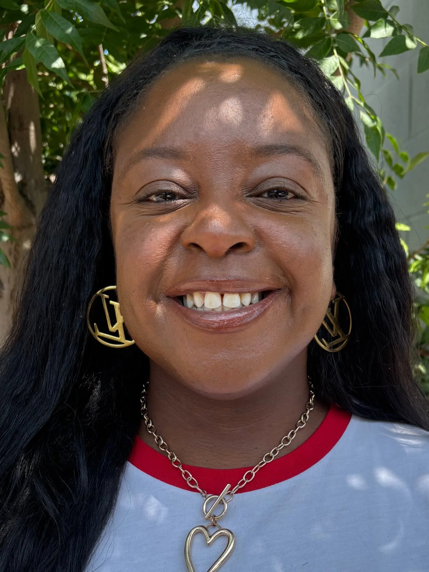 A smiling woman wearing a white-and-red shirt and hoop earrings standing outside next to a green tree