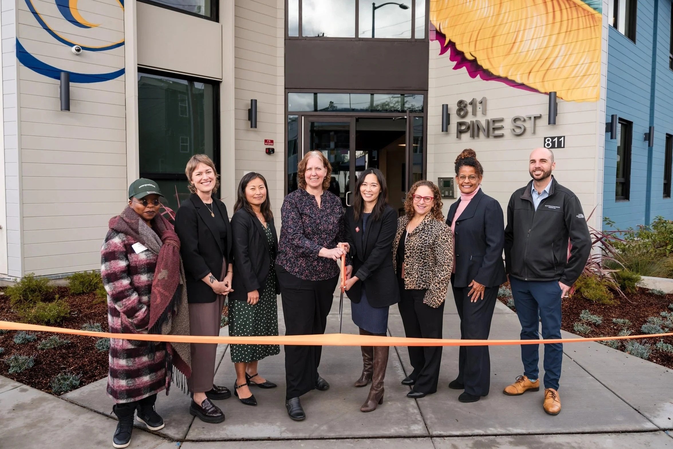 A group of smiling people hold large scissors while getting ready to cut an orange ribbon outside a new apartment building in Oakland