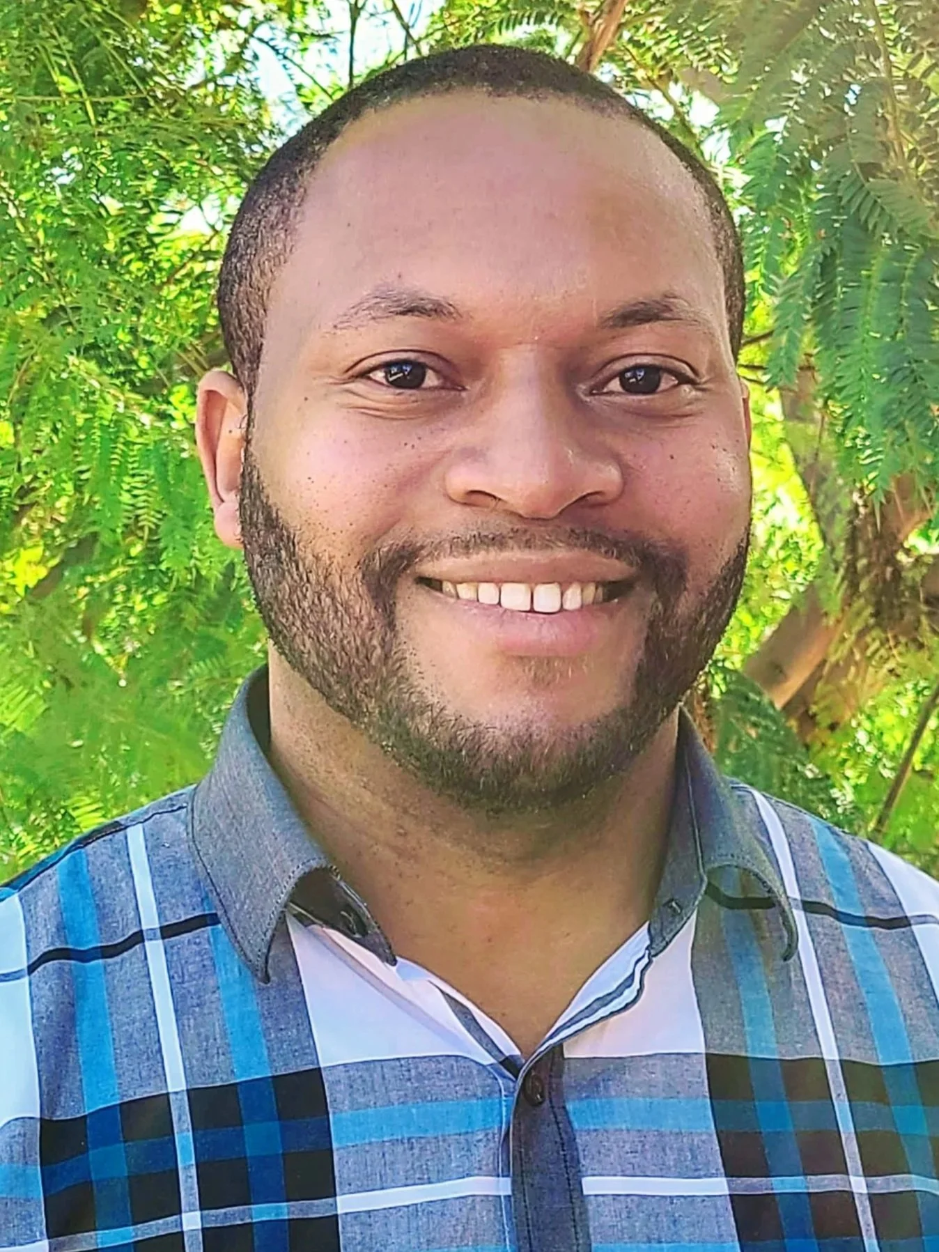 A bearded man smiling and wearing a blue, plaid collared shirt while standing outside in front of a green tree