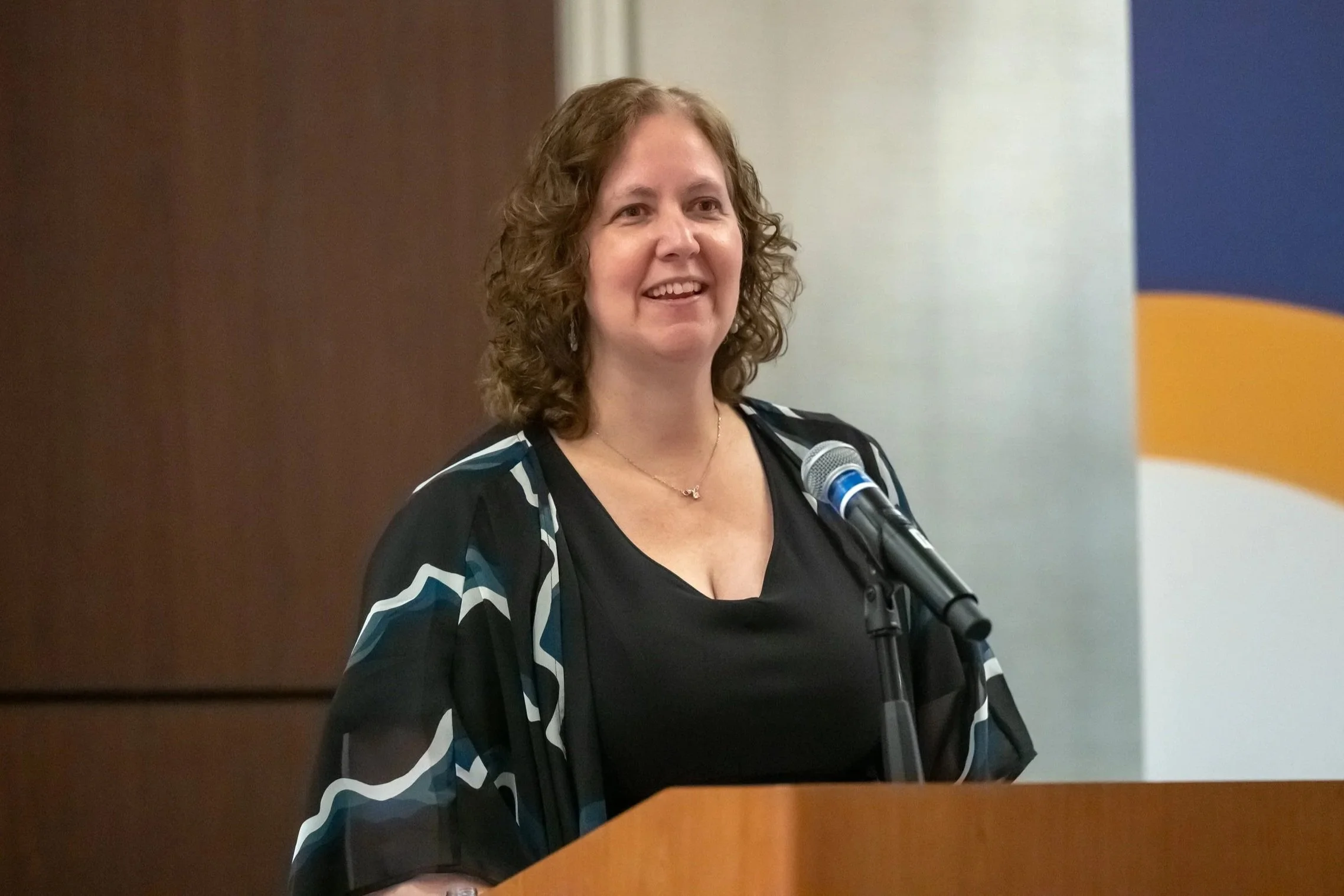 A smiling woman wearing a dark blouse delivers a speech inside a large ballroom
