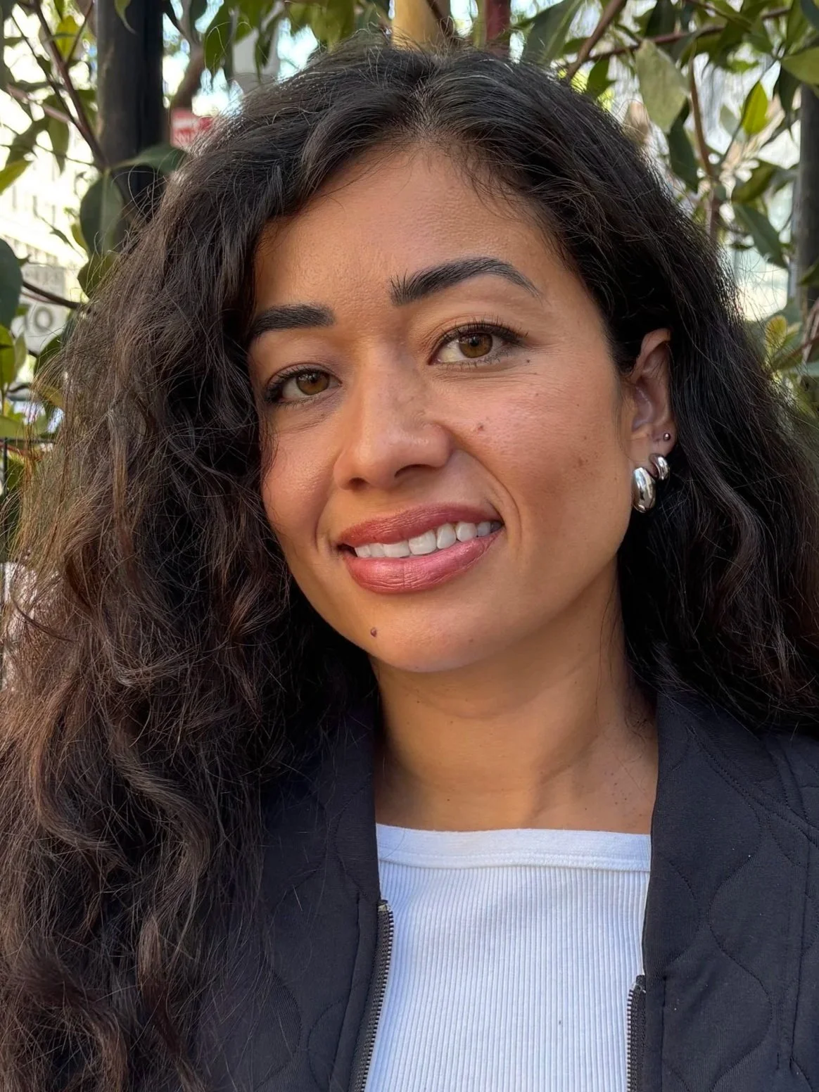 A woman wearing a dark jacket and a light blue blouse standing outside in front of a tree on an overcast day