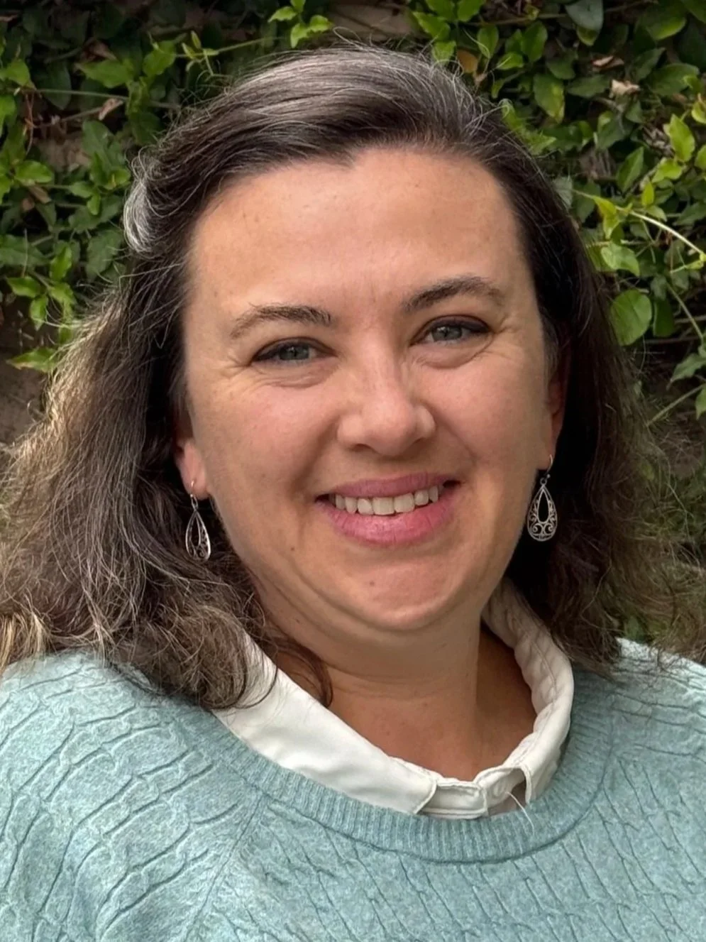 A smiling woman wearing a light green sweater and a white collared shirt posing for a photo while standing in front of shrubbery