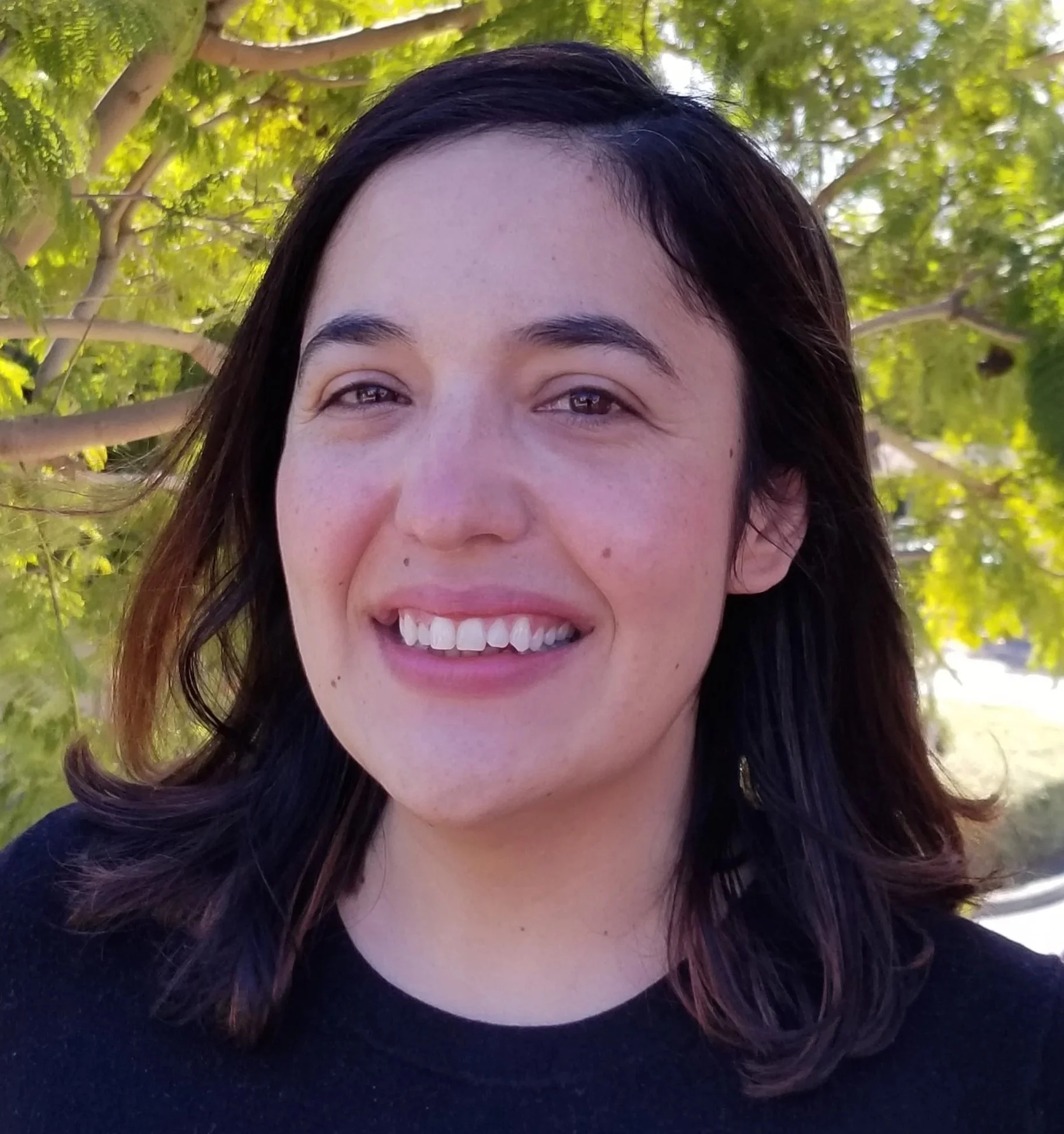 A smiling woman wearing a black blouse standing in front of a green tree on a sunny day