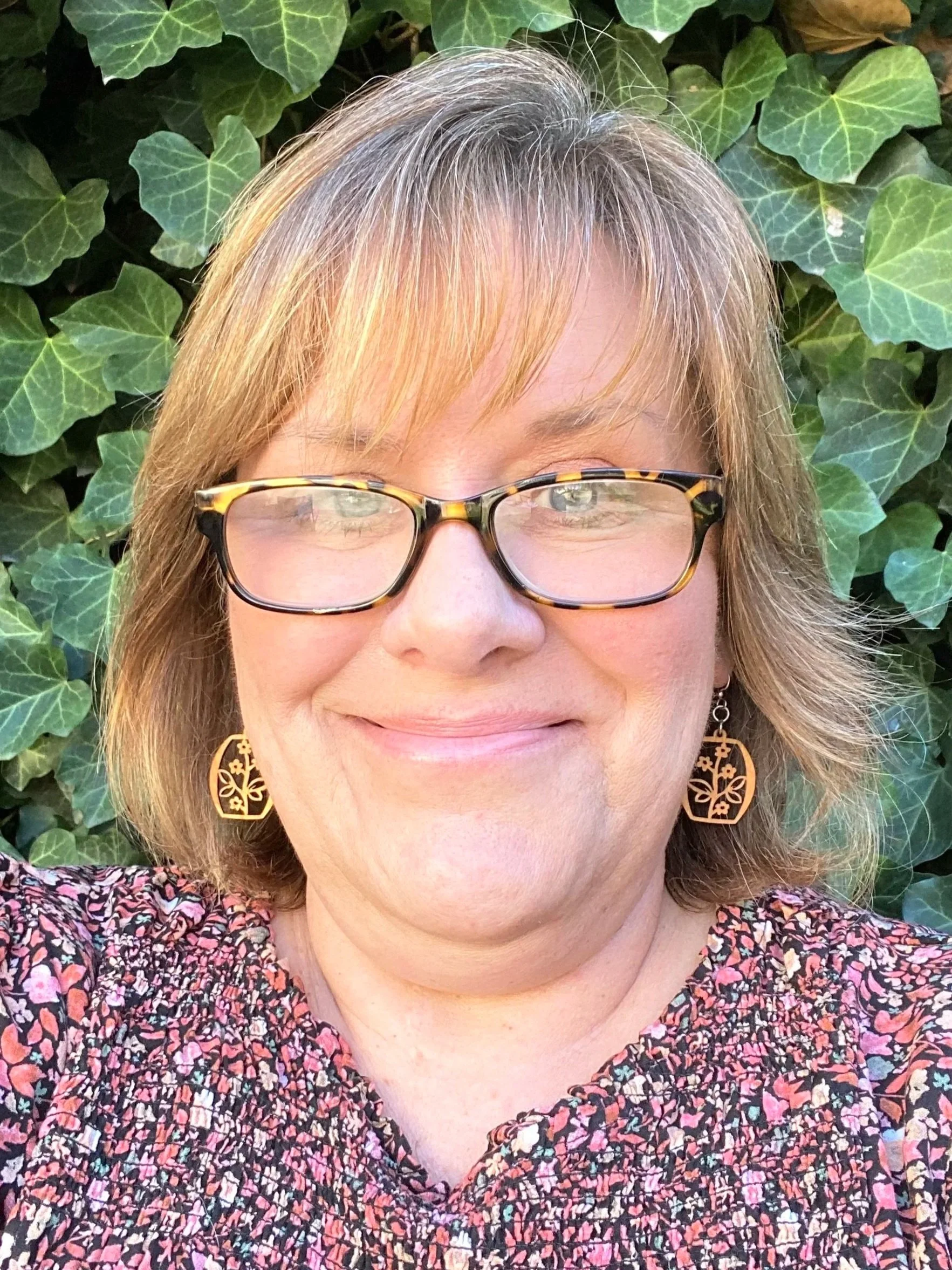 A woman wearing glasses and a black-and-red blouse standing in front of green ivy