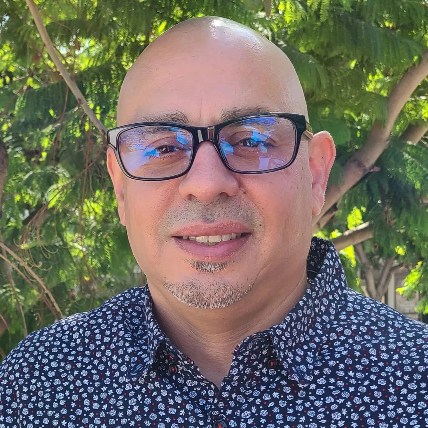 A man in a blue dress shirt and glasses standing in front of a tree
