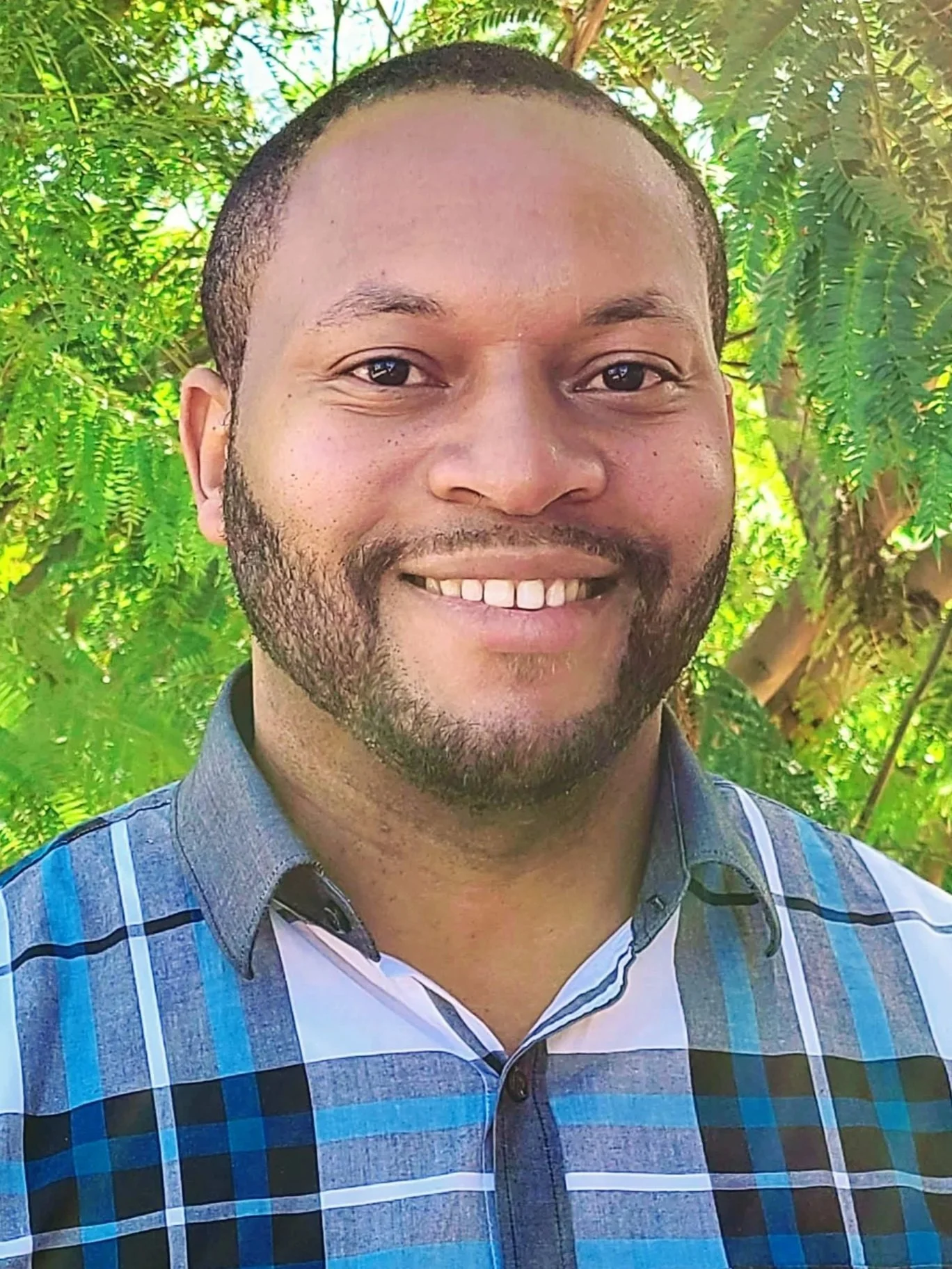 A bearded man smiling and wearing a blue, plaid collared shirt while standing outside in front of a green tree