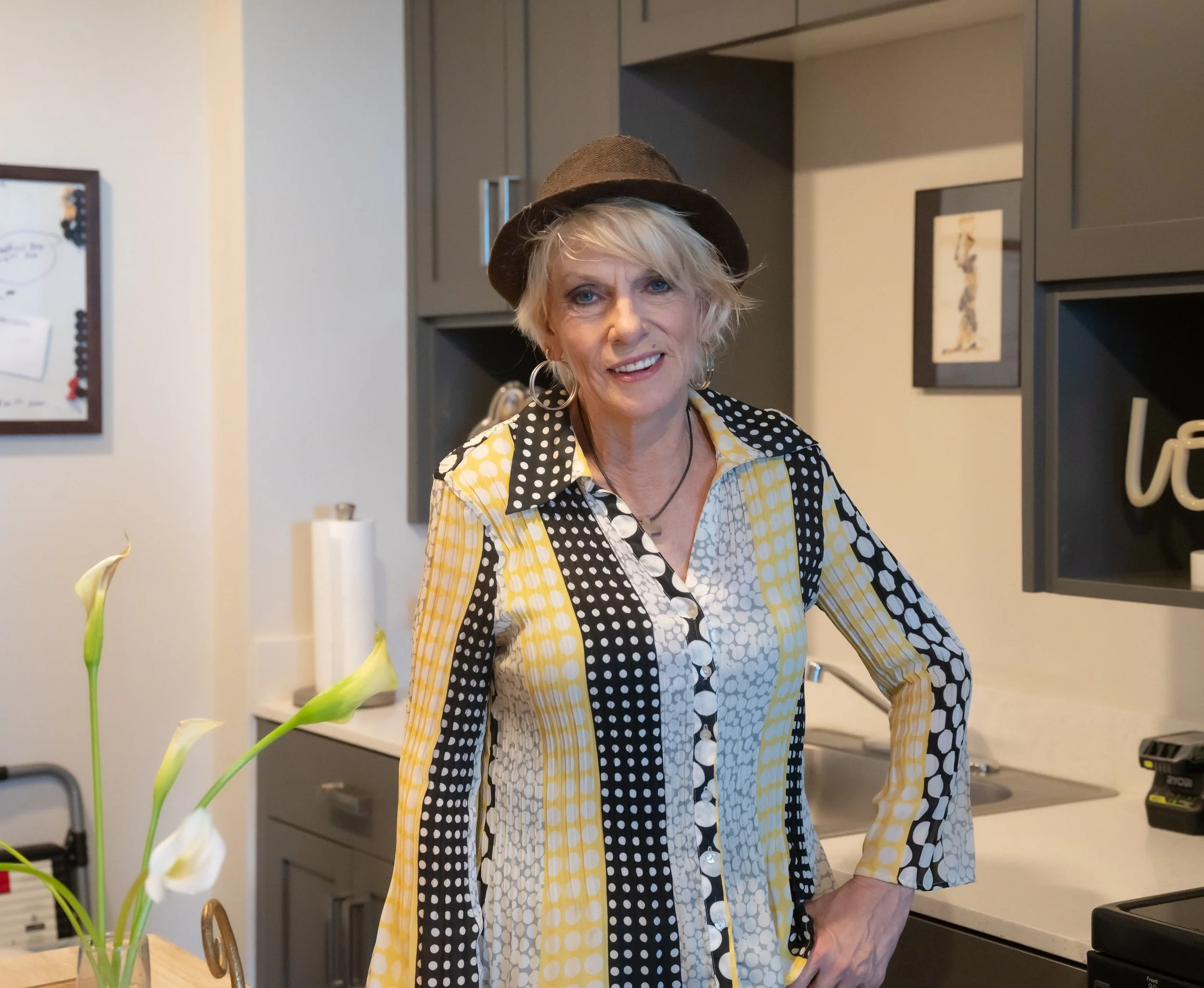 A smiling woma wearing a multi-color blouse and brown fedora hat standing in her new kitchen
