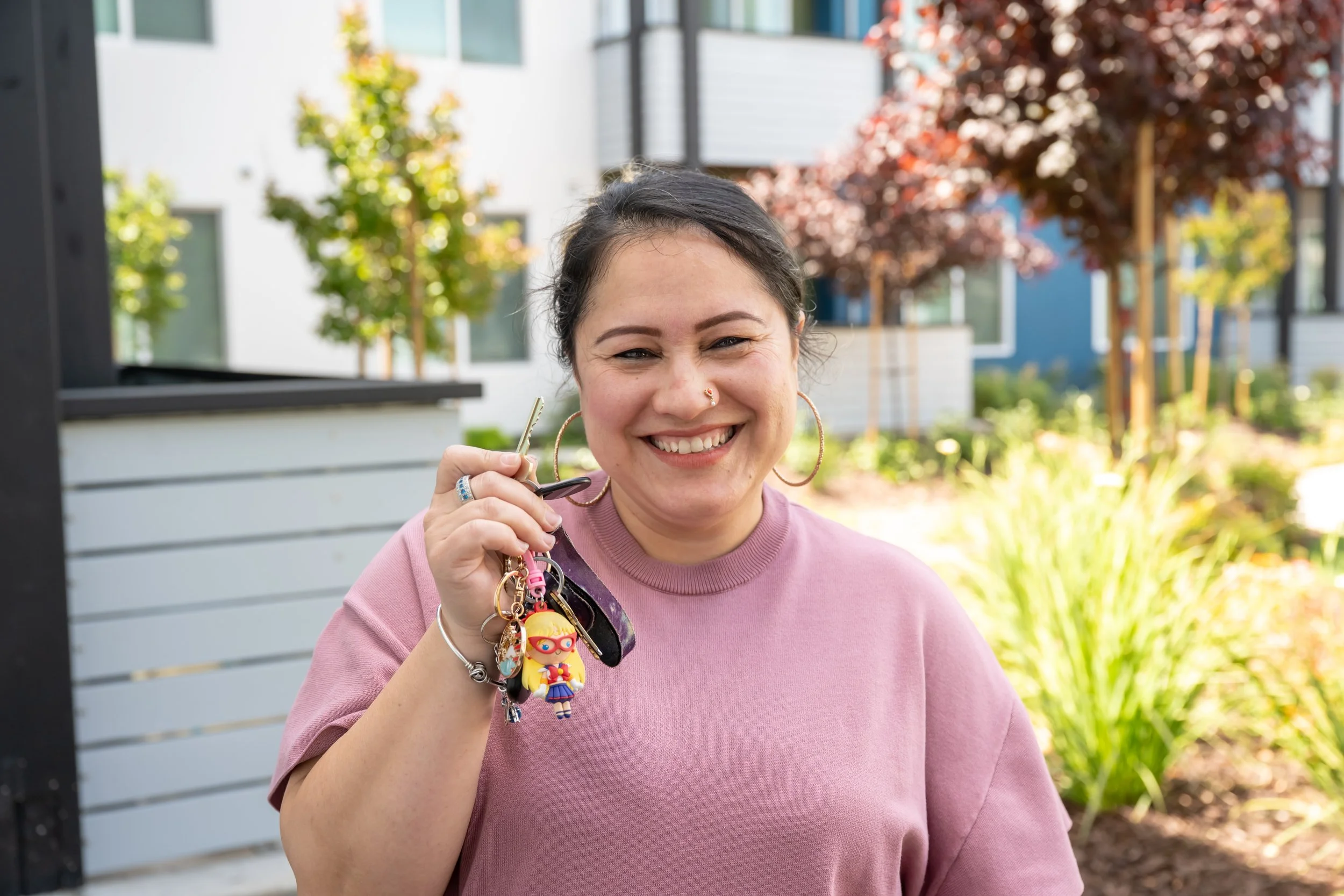 A smiling woman wearing a pink sweatshirt and holding a house key standing in front of a white apartment building on a sunny day