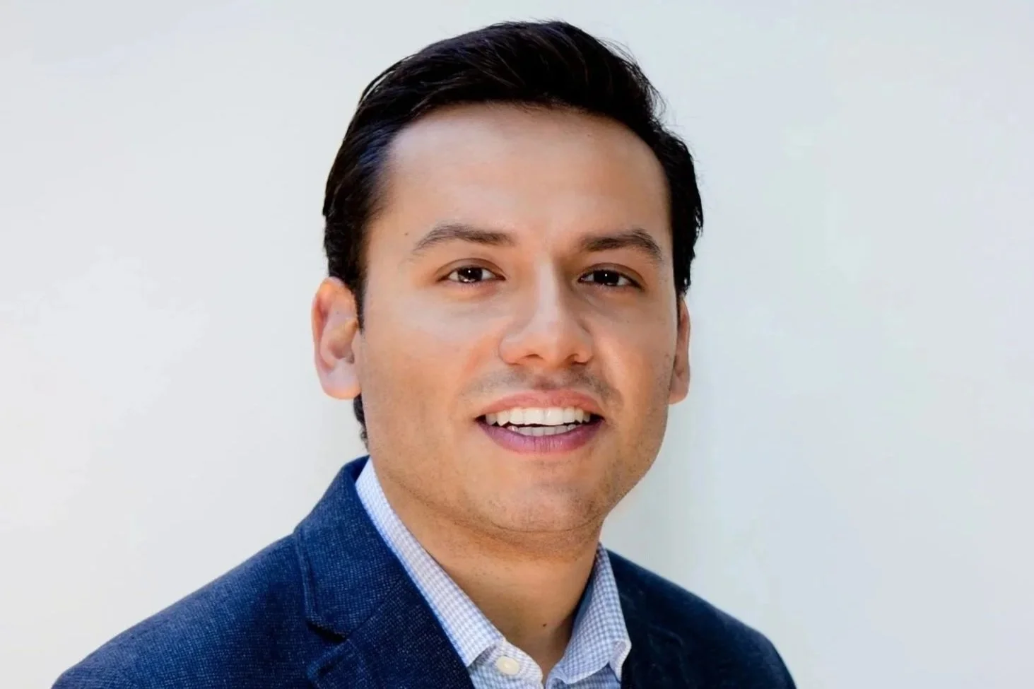 A smiling man in a blue blazer and a dress shirt poses for a photo in front of white-color wall