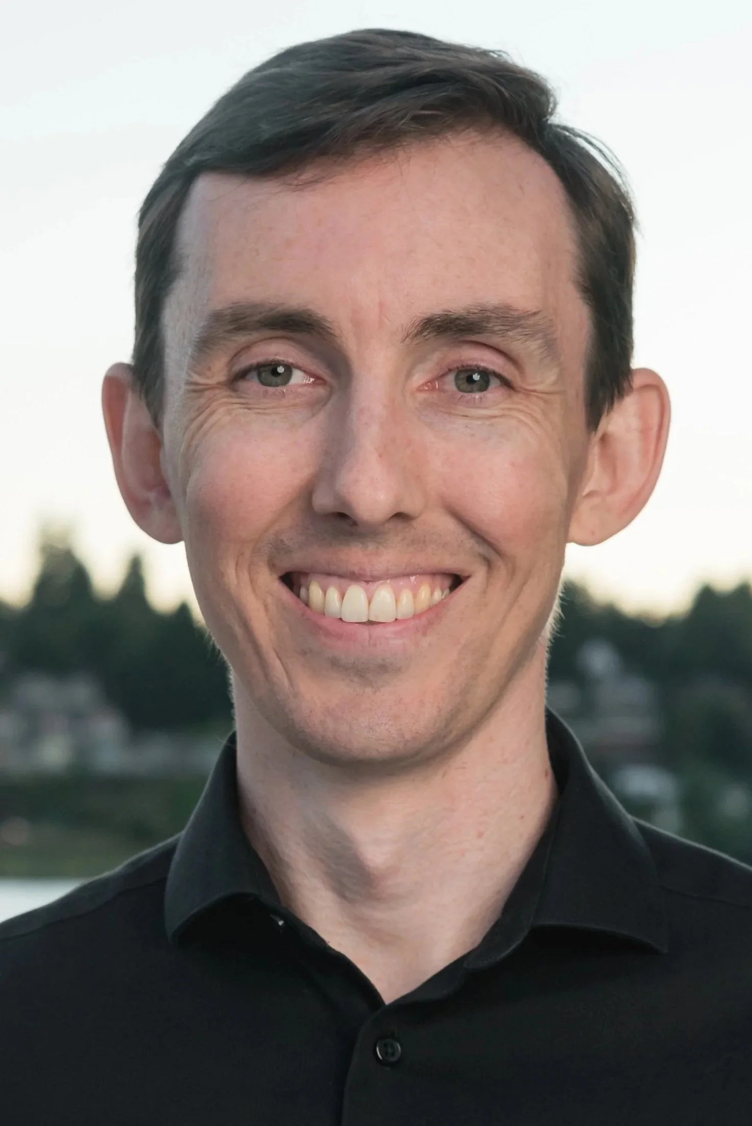 A smiling man wearing a black dress shirt standing outside at twilight