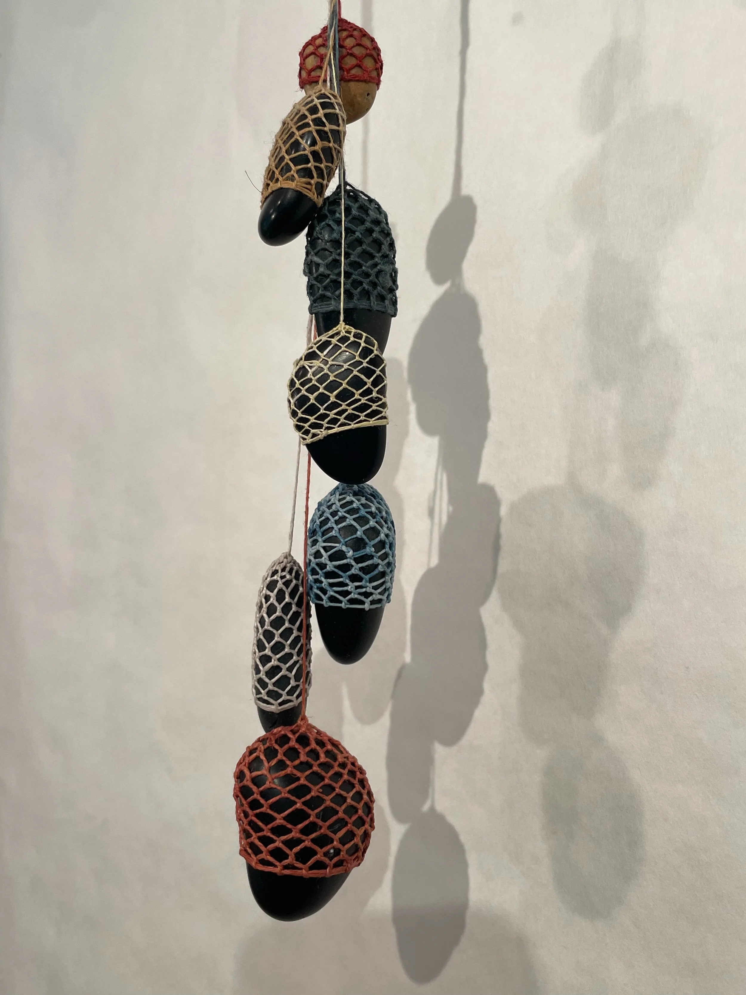 Building on the Buddhist metaphor of Indra's net, this series suspends several small netted stones from a cast-iron hook - tumbling down in a cascade of stones.
Maine beach stones with naturally dyed waxed Linen. 
Approximately 12" by 2" by