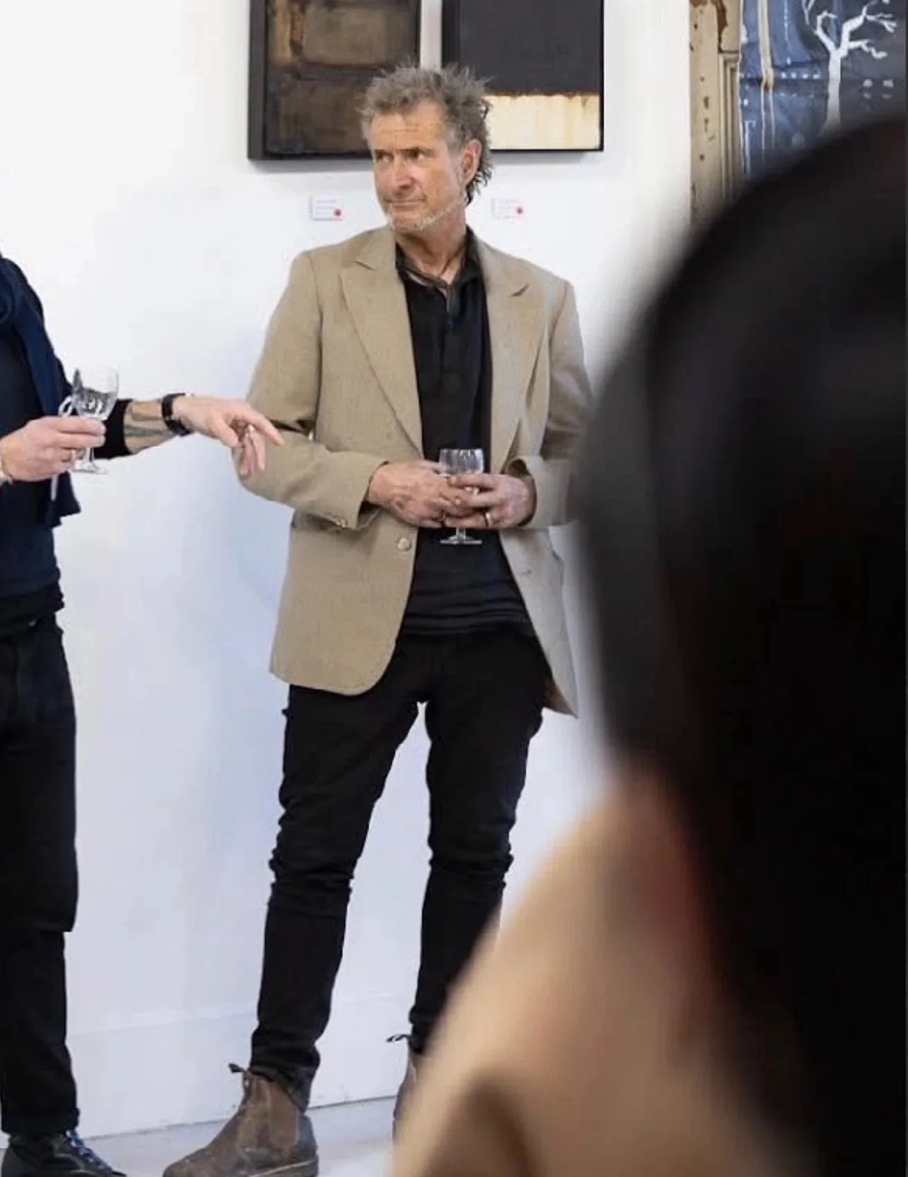A man with gray hair and a beard, wearing a beige blazer, black shirt, and dark pants, is holding a glass and looking to his right during a social event.