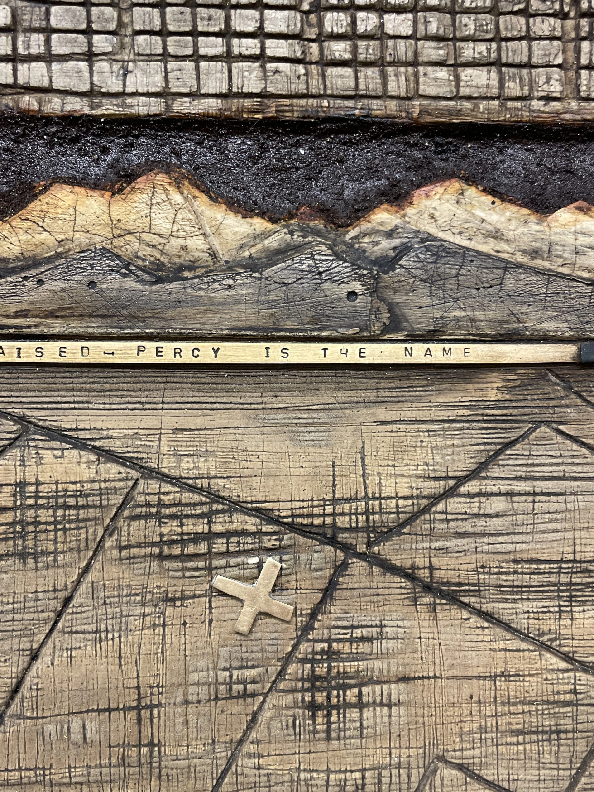 Close-up of a wooden surface with carved details, a metal strip with the inscription 'PERCY IS THE NAME,' a small metal cross, and a painted mountain range in the background.