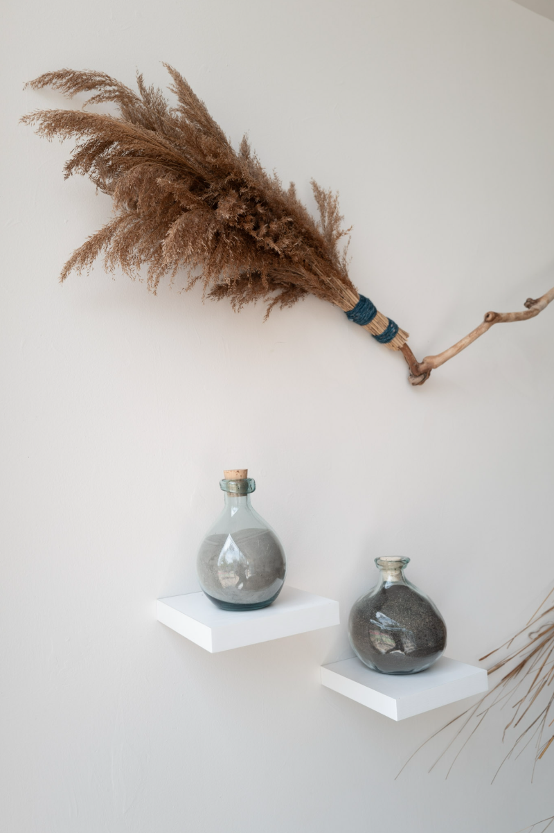 Detail of a handmade broom made from dried reeds and a branching wooden handle above two glass bottles filled with gray volcanic ash.